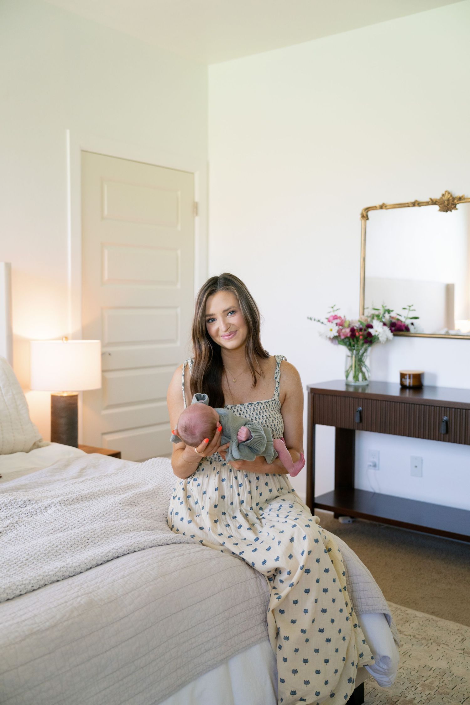 Young woman in polka dot dress sitting on bed holding decorative items in a bright, elegant bedroom.