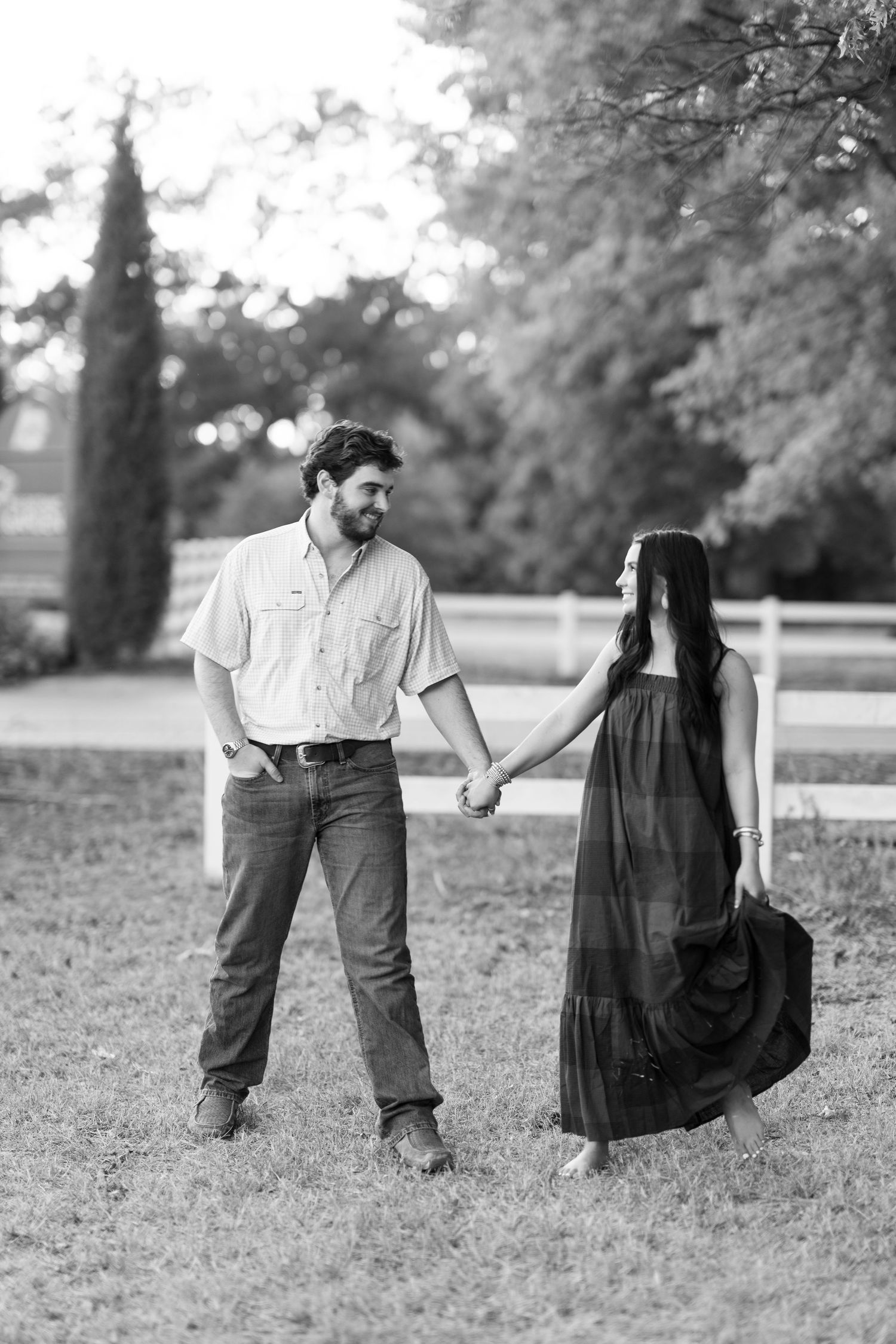 Black and white photo of a couple holding hands walking in a grassy field near a white fence.
