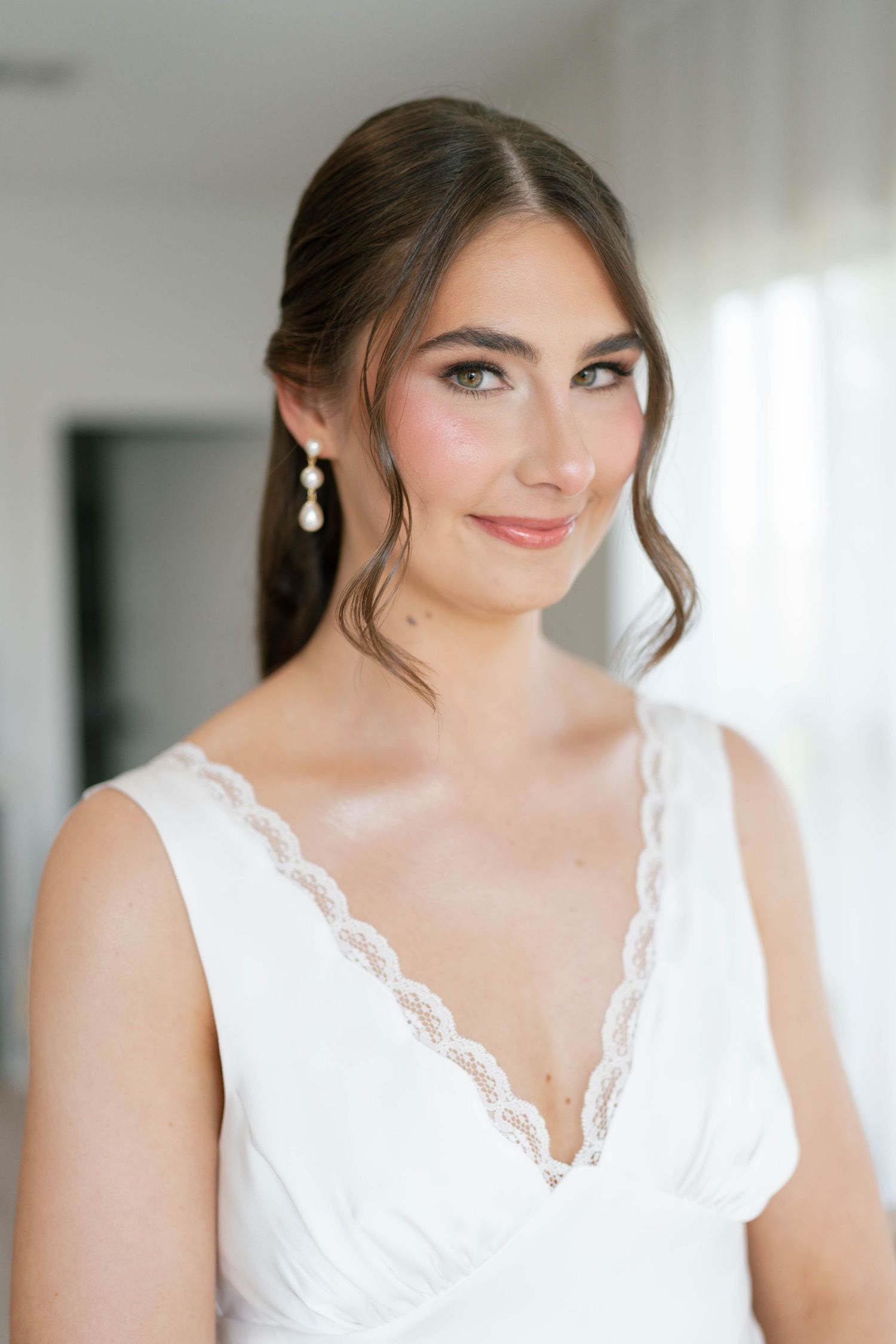 Elegant bride in a white lace gown wearing pearl drop earrings smiling softly in a bright indoor setting.