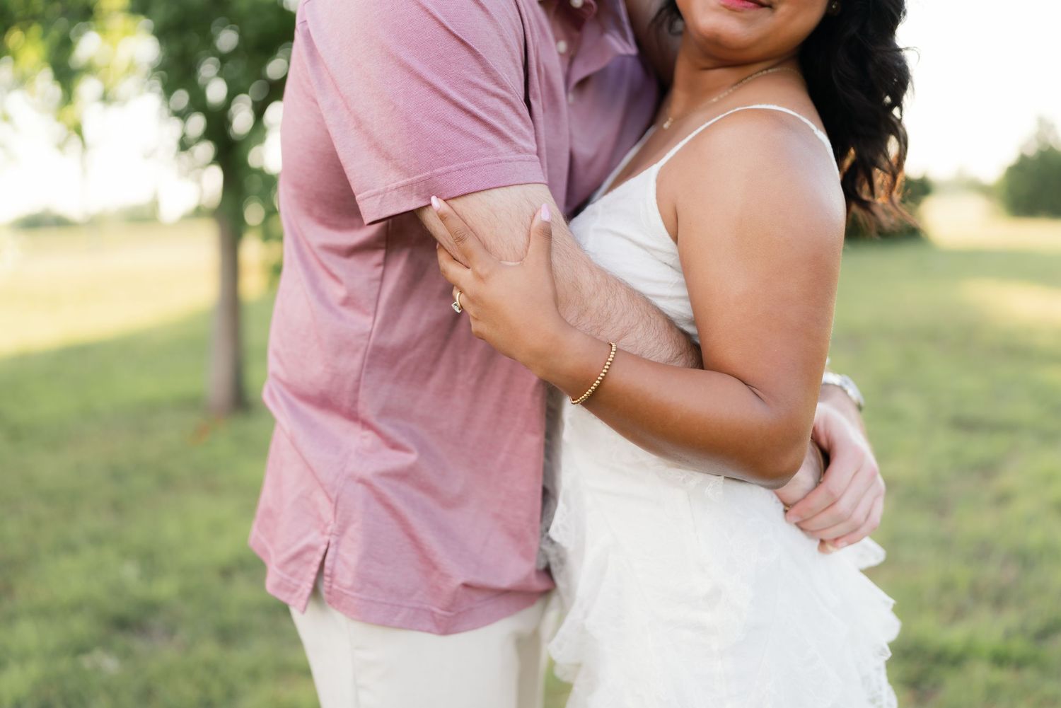 Couple embracing outdoors in a sunny park, man in pink shirt and woman in white dress.