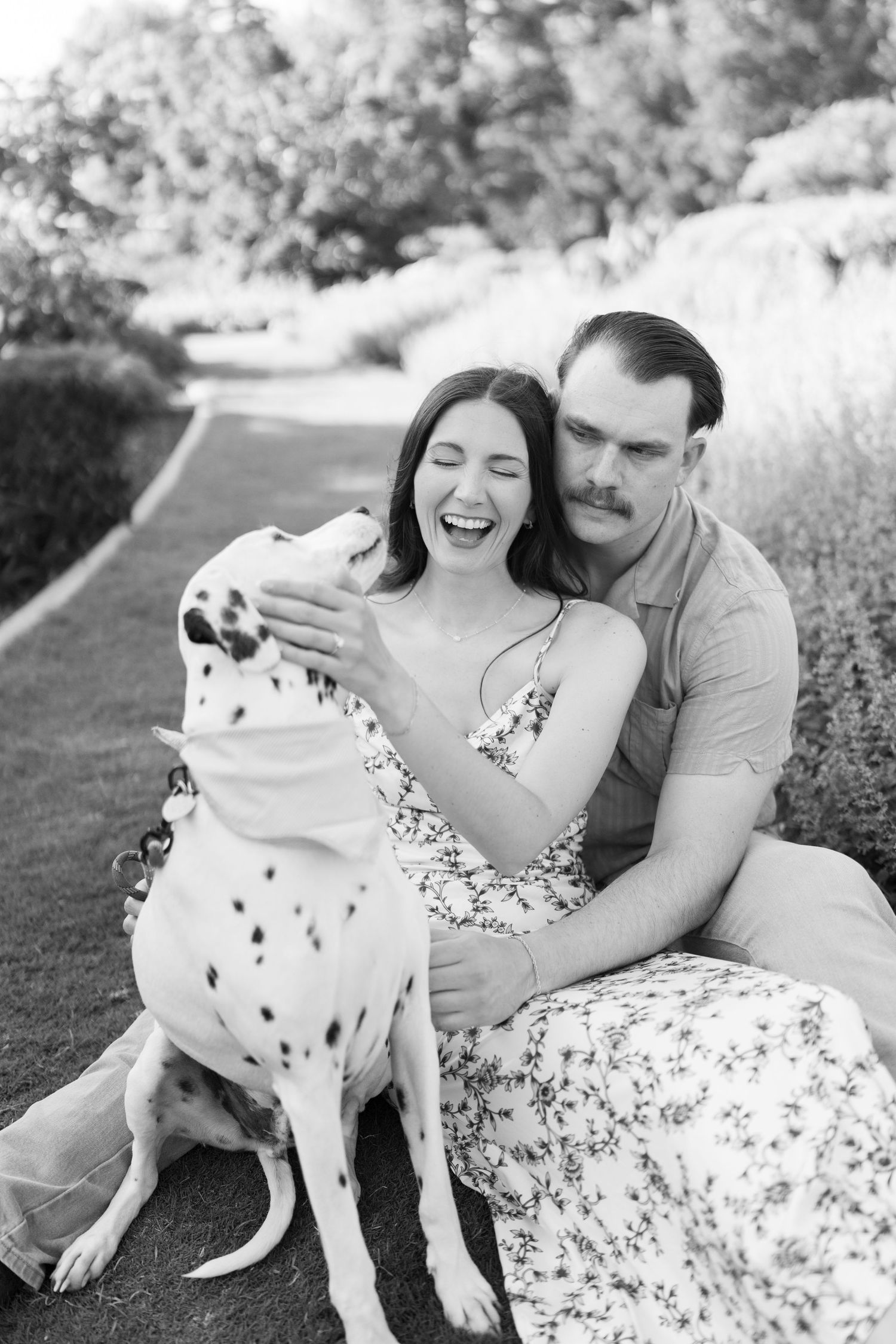 Black and white photo of a laughing couple sitting outdoors with their Dalmatian dog in a lush garden setting.