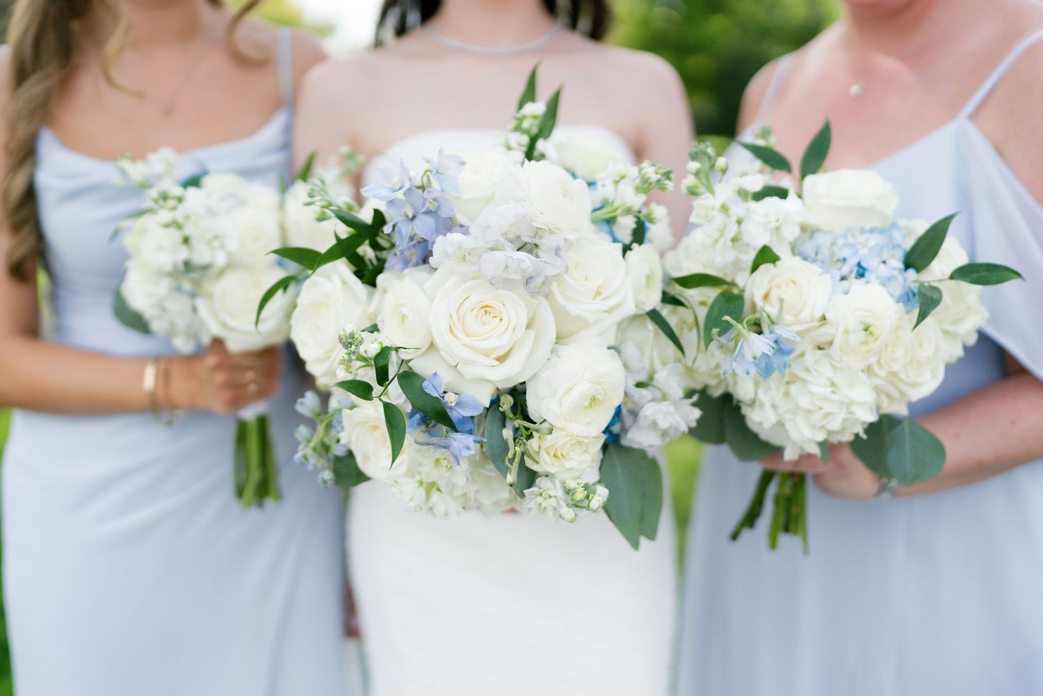 Bride and bridesmaids holding elegant white and blue floral bouquets with roses and hydrangeas at a wedding.