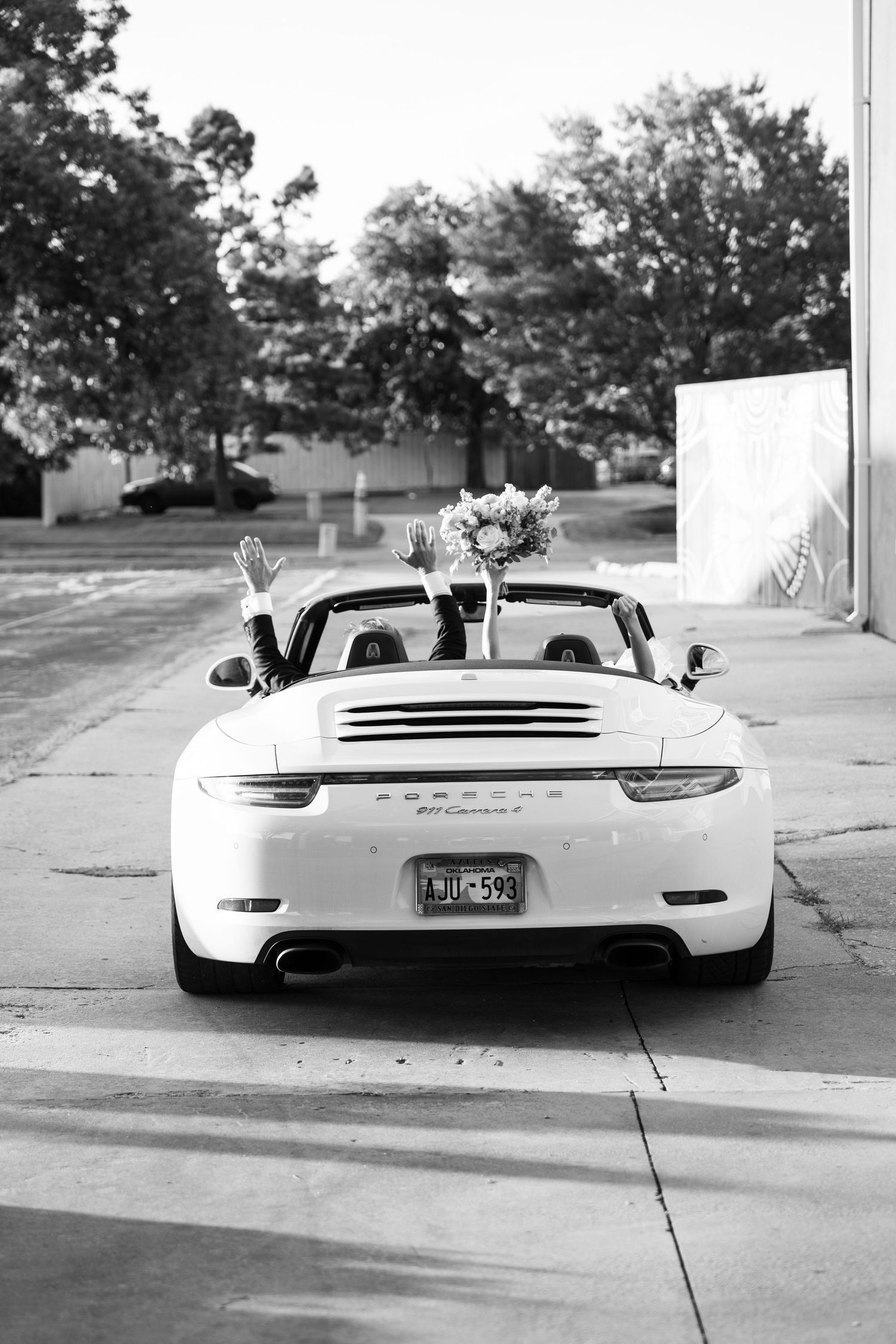 Black and white photo of white Porsche 911 Carrera convertible with wedding bouquet at a venue.