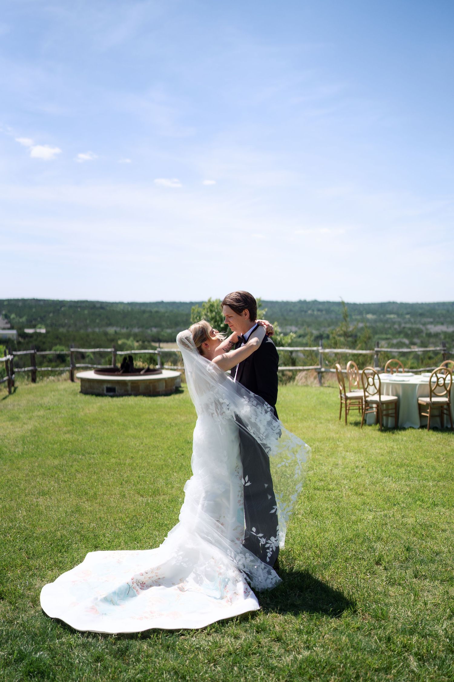 Two brides share a romantic dance outdoors on a sunny wedding day, with a flowing veil and scenic green landscape behind them.