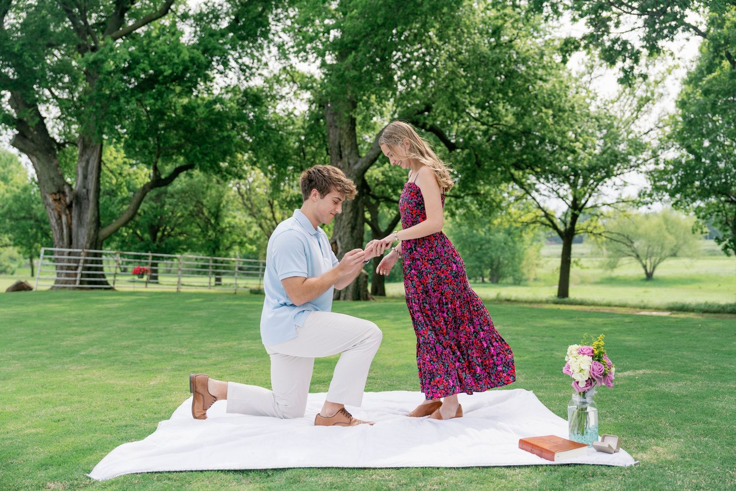 Man on one knee proposing to woman in floral dress on a white blanket in a lush green park.
