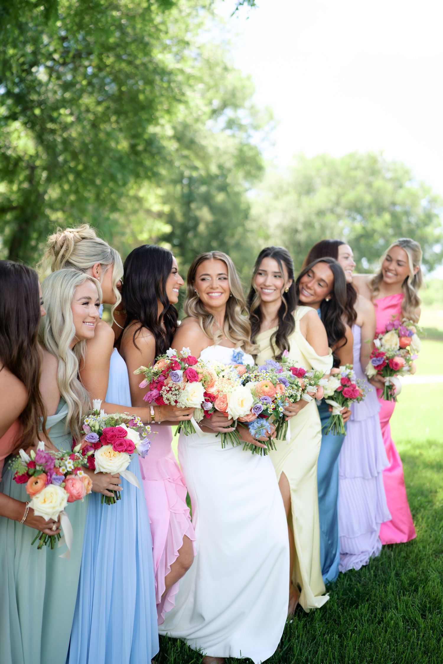 Bride surrounded by bridesmaids in colorful pastel gowns holding vibrant floral bouquets outdoors.