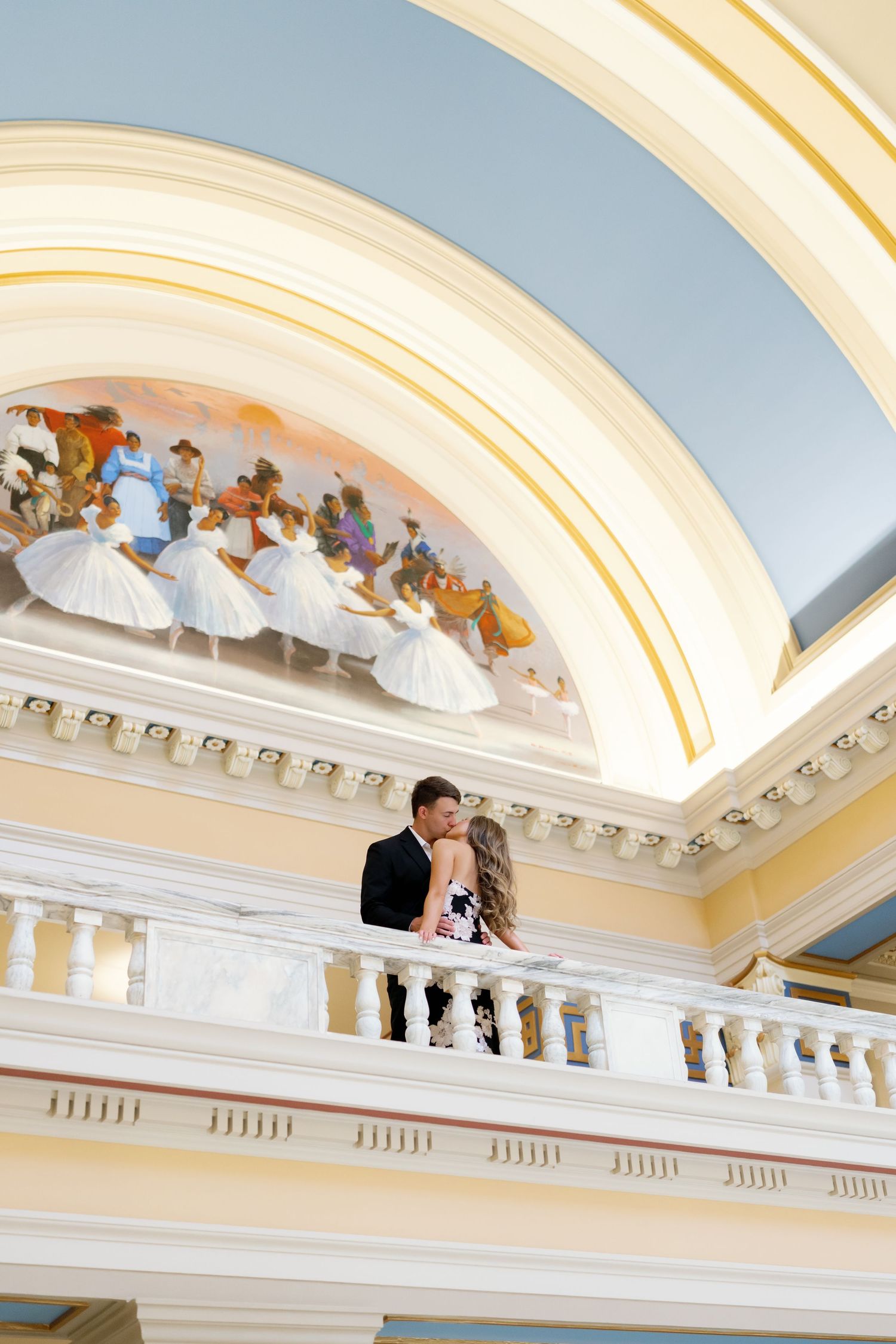Couple sharing a romantic kiss on an elegant balcony beneath a colorful mural in a grand historic building.