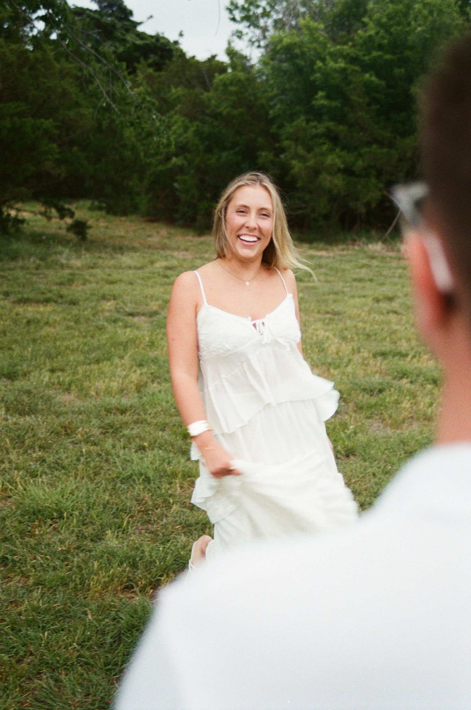 Smiling blonde woman in white sundress holding hands with partner in a lush green meadow.