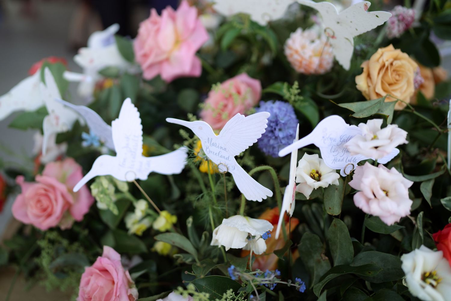 White hummingbird decorations accent a lush floral arrangement of pink roses and colorful blooms.