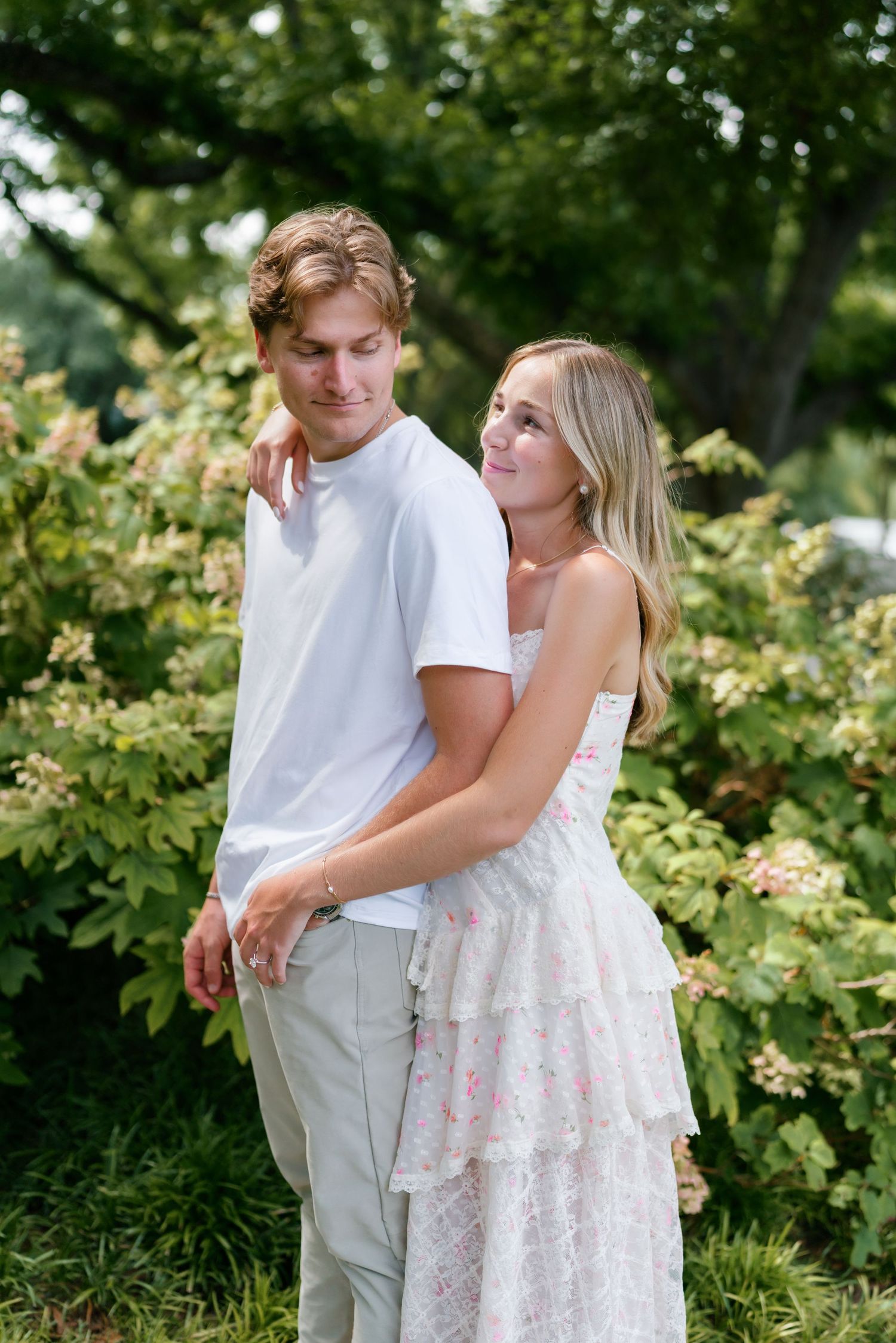 Couple embracing outdoors among lush green hydrangea bushes, woman in white floral dress smiling up at man in white tee.