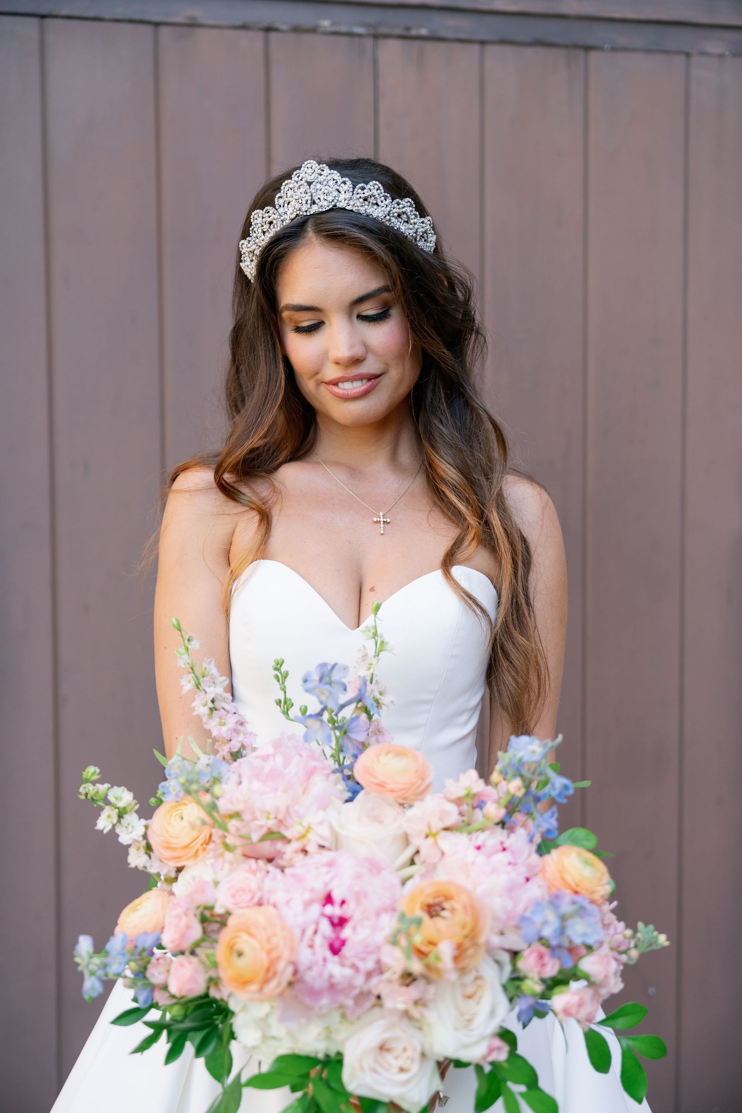 Beautiful bride in strapless white gown wearing tiara holding large pastel floral bouquet with roses.