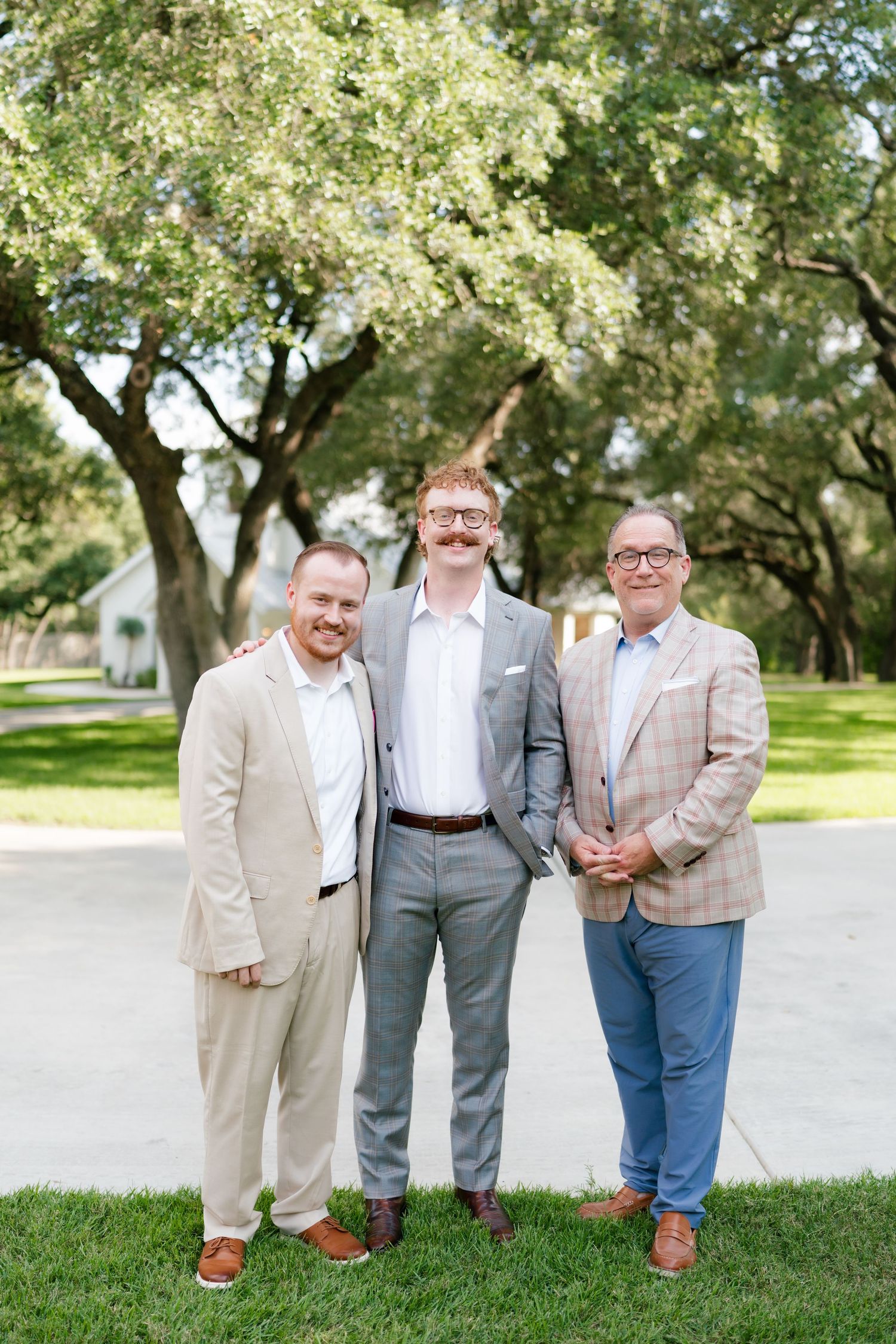 Three men in suits smiling together outdoors under large oak trees on a sunny day.