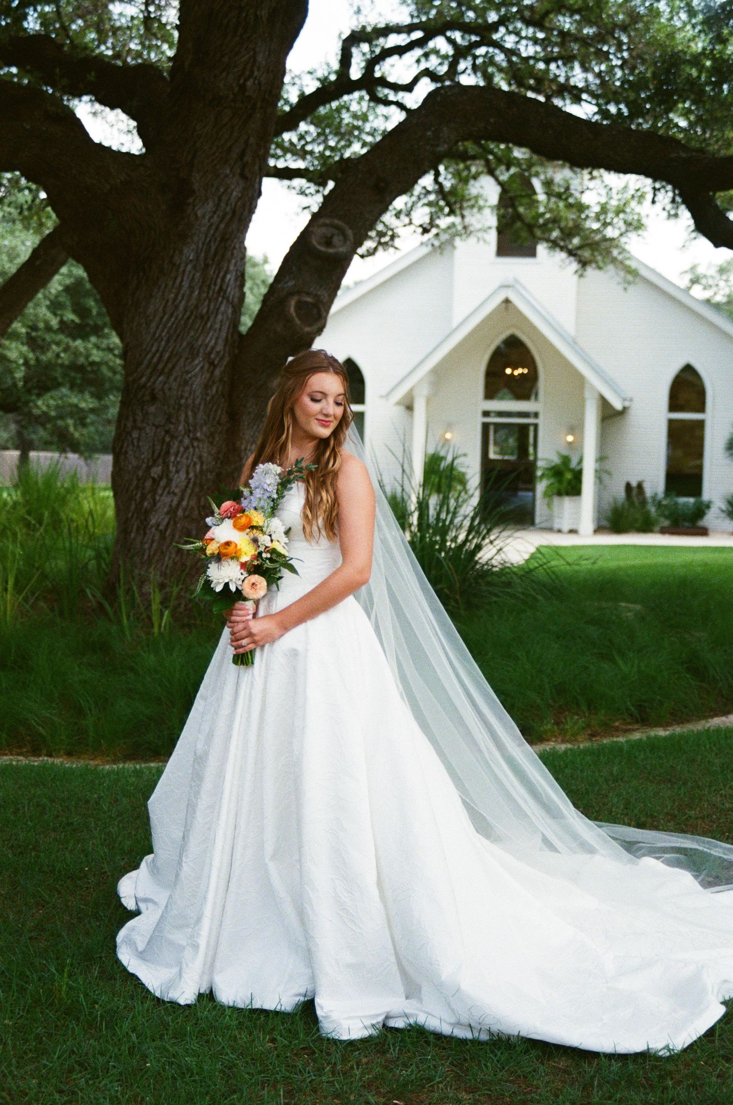 Elegant bride in ball gown with long veil holds bouquet beside oak tree near white chapel at dusk.