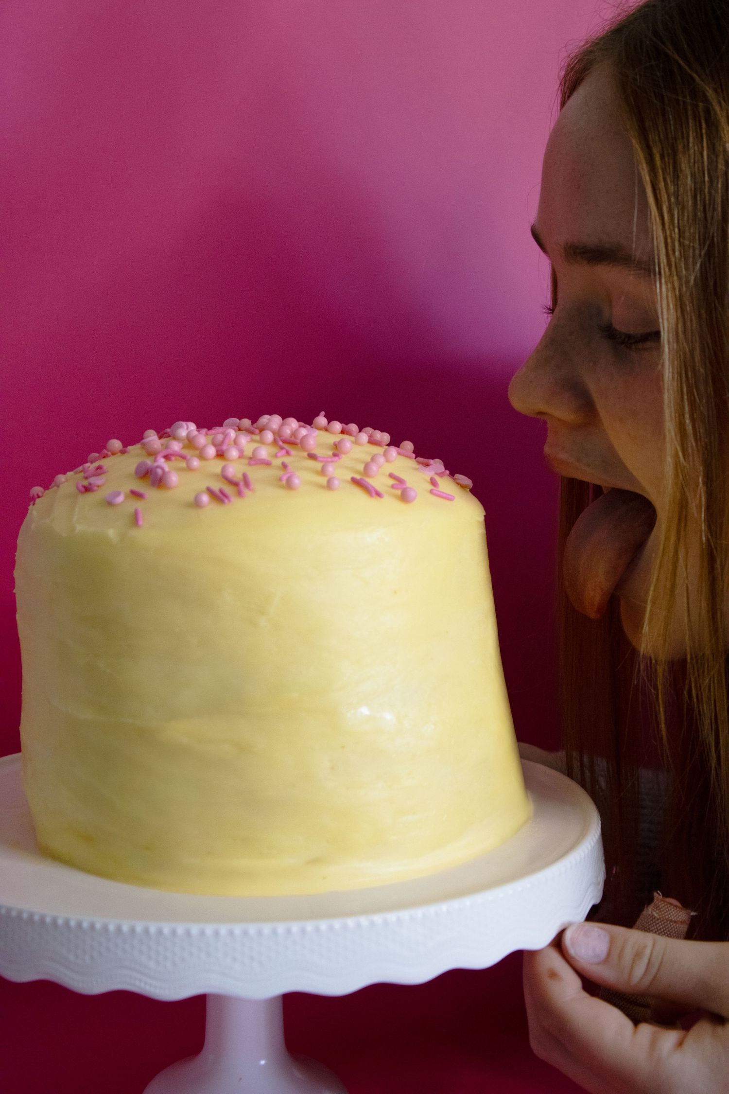 Young woman admiring a yellow frosted birthday cake with pink sprinkles on a white cake stand against pink background.