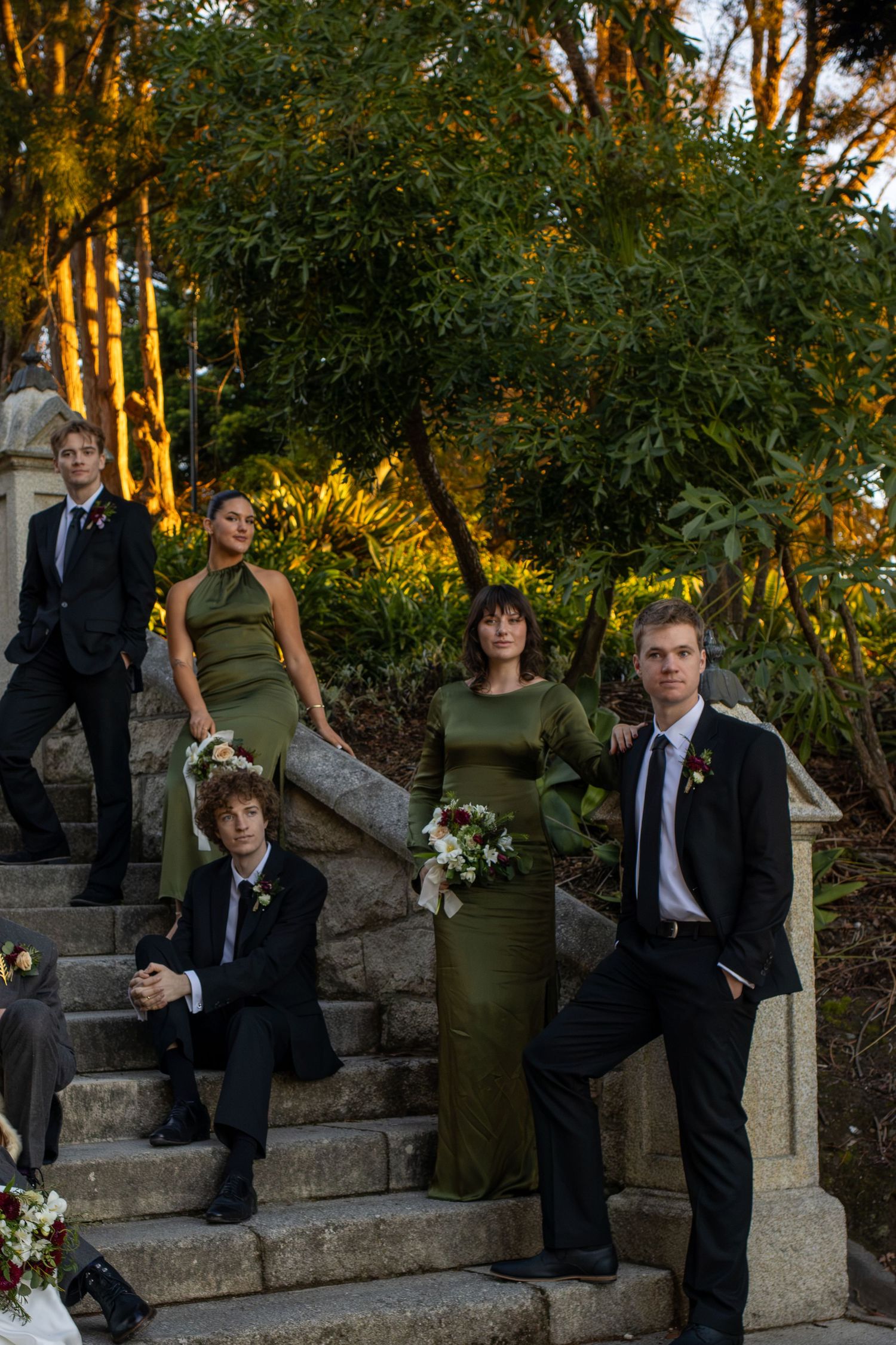 Wedding party in olive green dresses and black suits posing on stone steps at sunset.