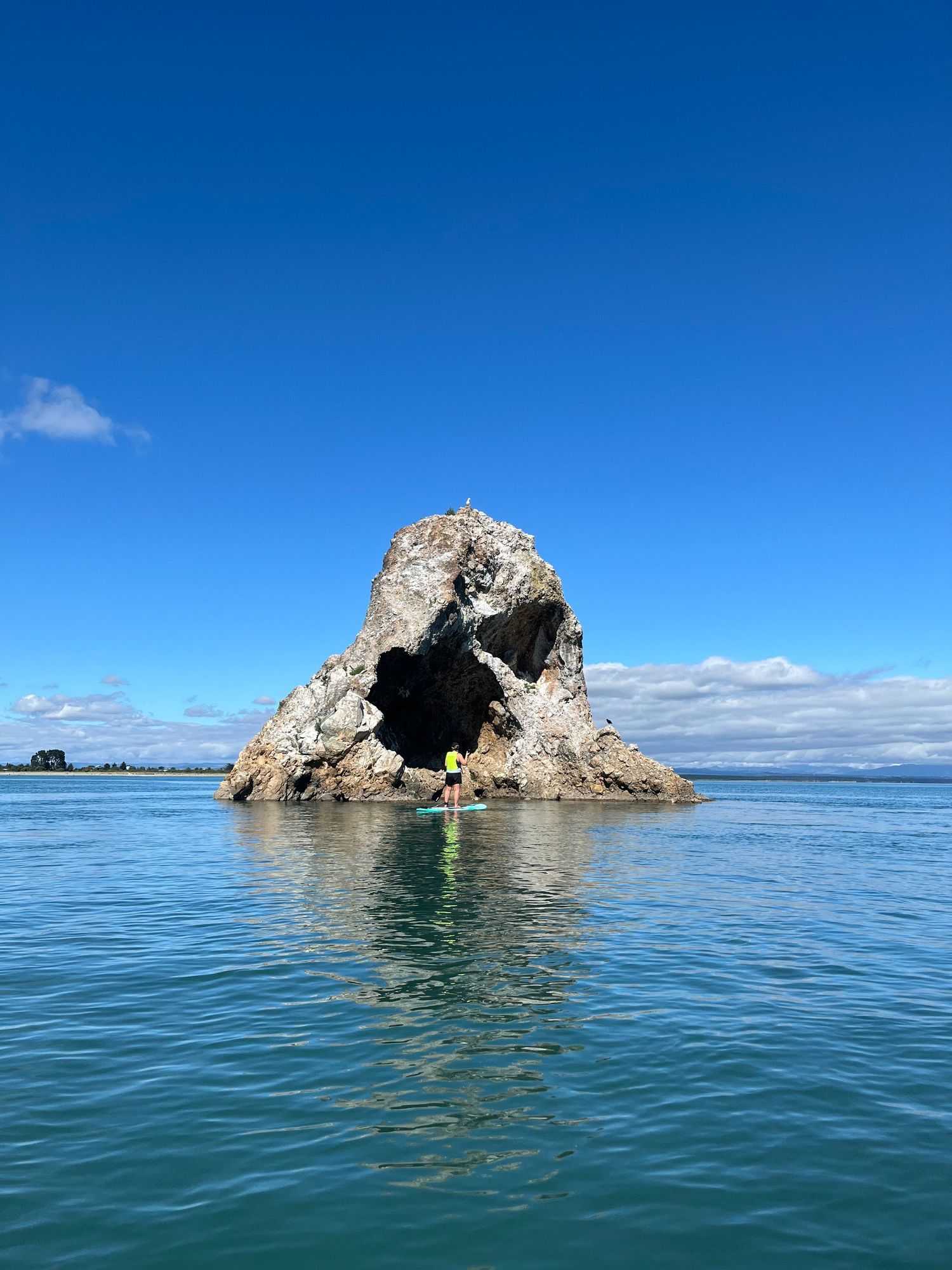 Person on paddleboard approaching dramatic rock arch formation in turquoise ocean water.