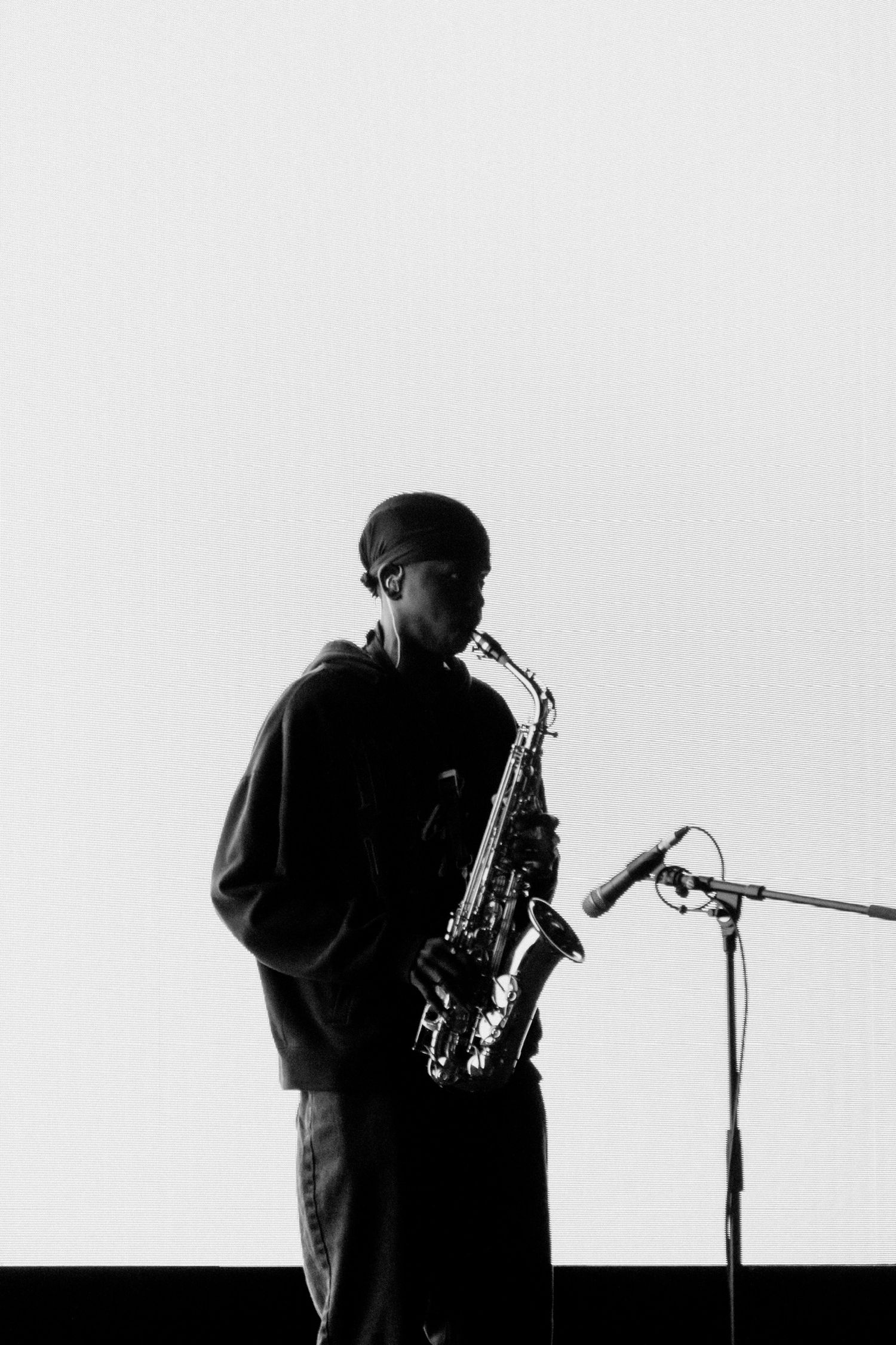 Black and white photo of musician playing saxophone on stage near microphone.