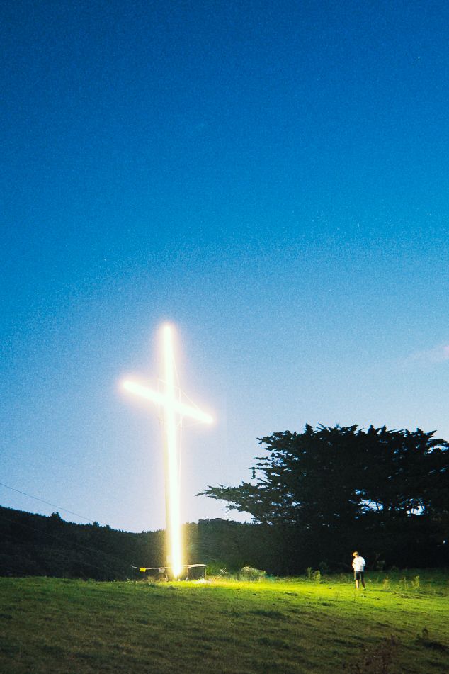 Bright illuminated cross glowing against a deep blue twilight sky with a silhouetted tree nearby.