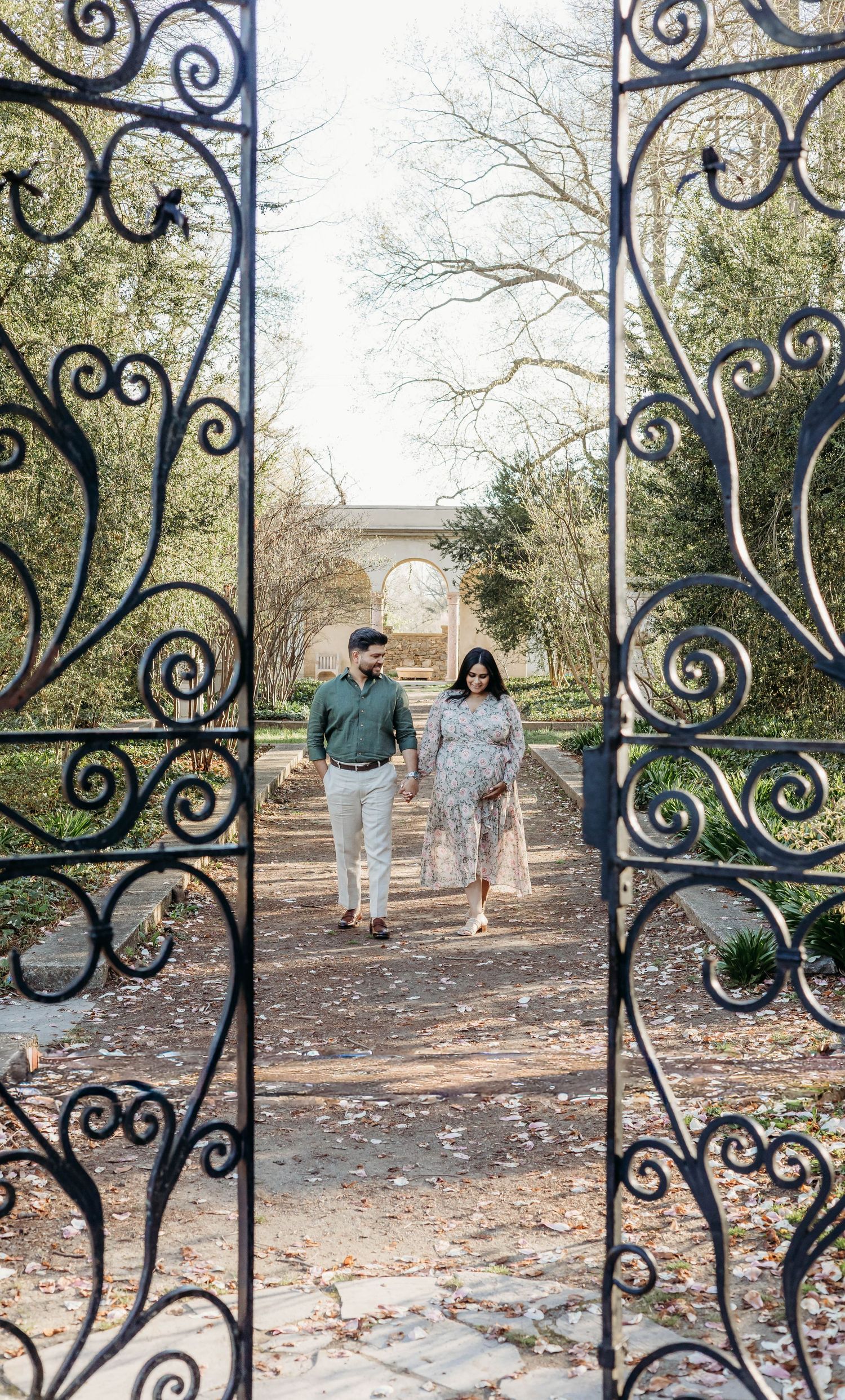Expecting couple walks hand-in-hand through ornate iron garden gates during a maternity photography session.