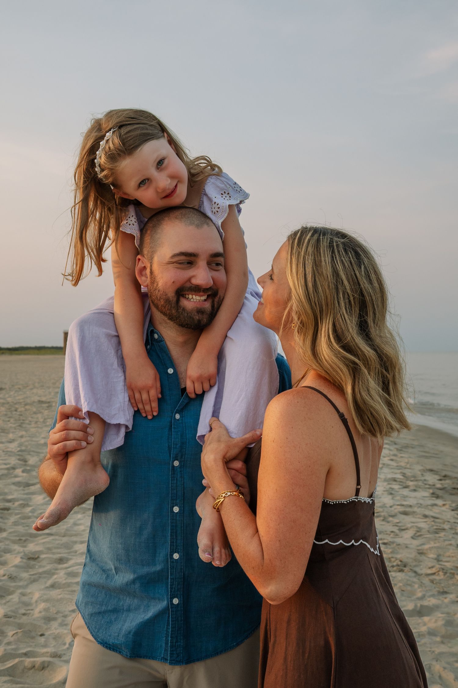 A happy family of three enjoys a beach sunset, with a smiling girl on her father's shoulders as her mother looks on.