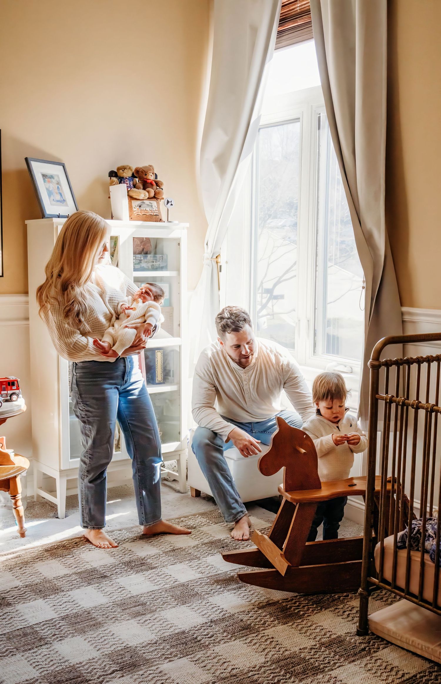Family with newborn and toddler enjoy a warm moment in a sunlit vintage-styled nursery room.