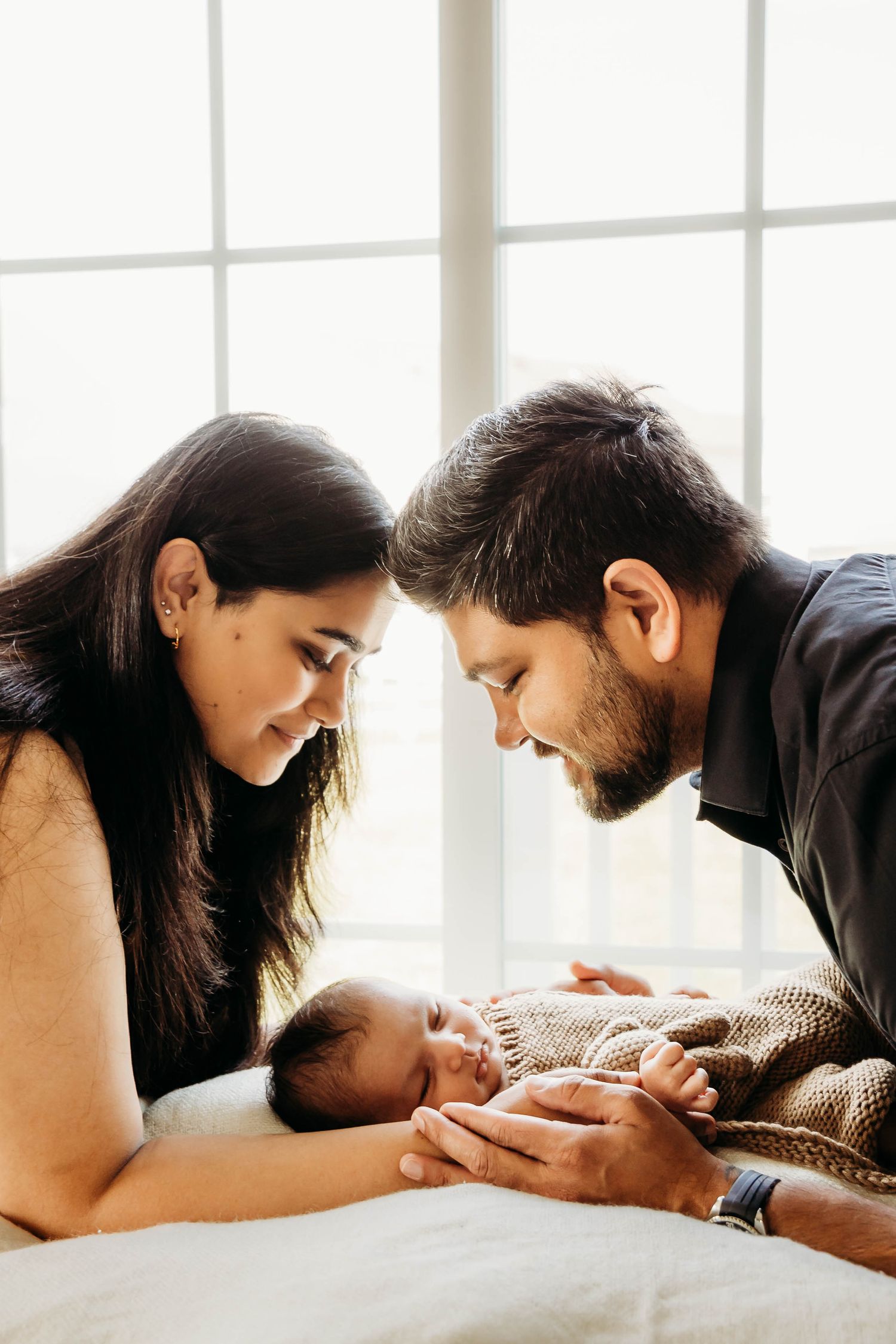 Parents leaning over their newborn baby lying on a white blanket in front of a bright window.