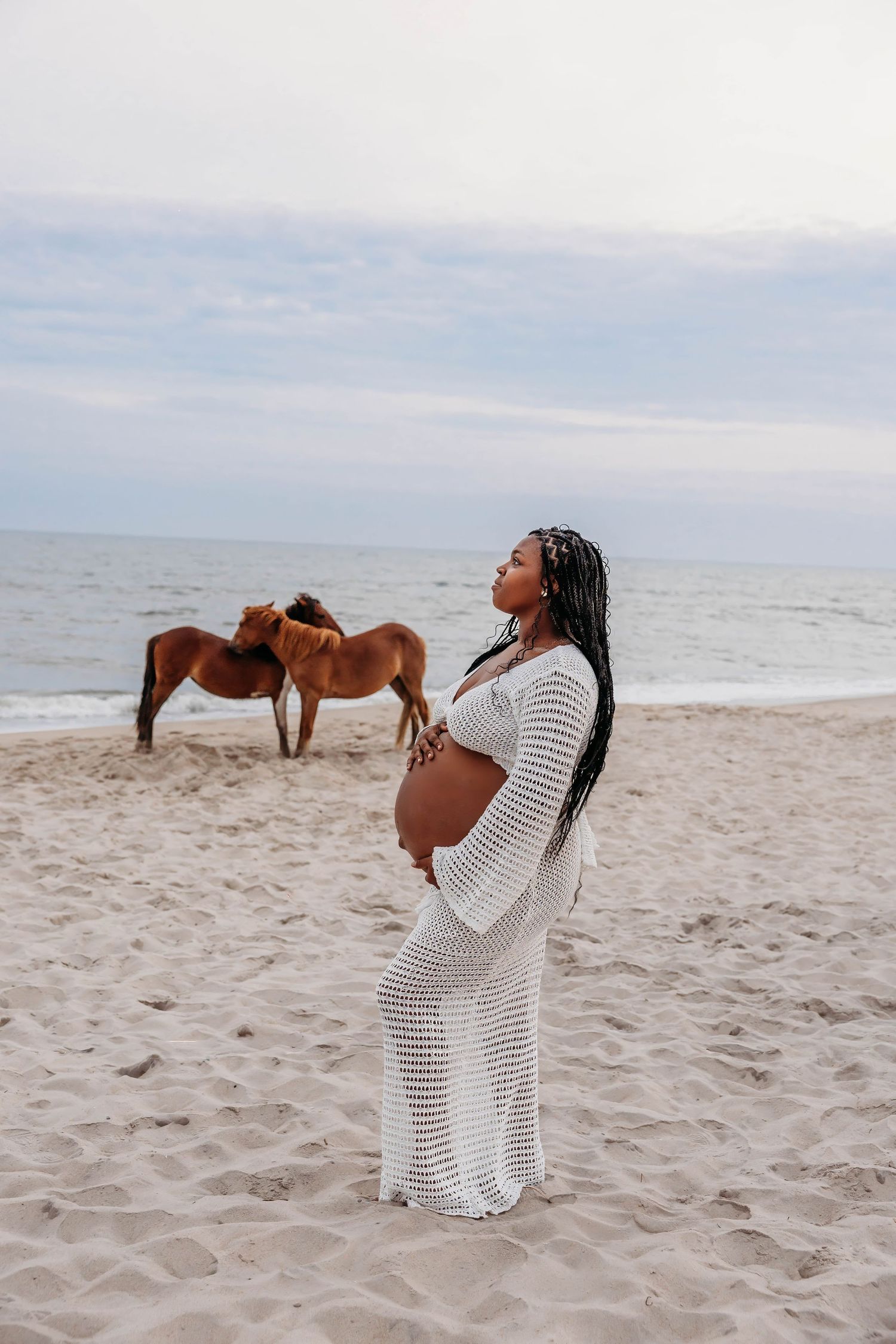 A pregnant woman in a white crochet dress stands on a beach with wild horses grazing behind her.
