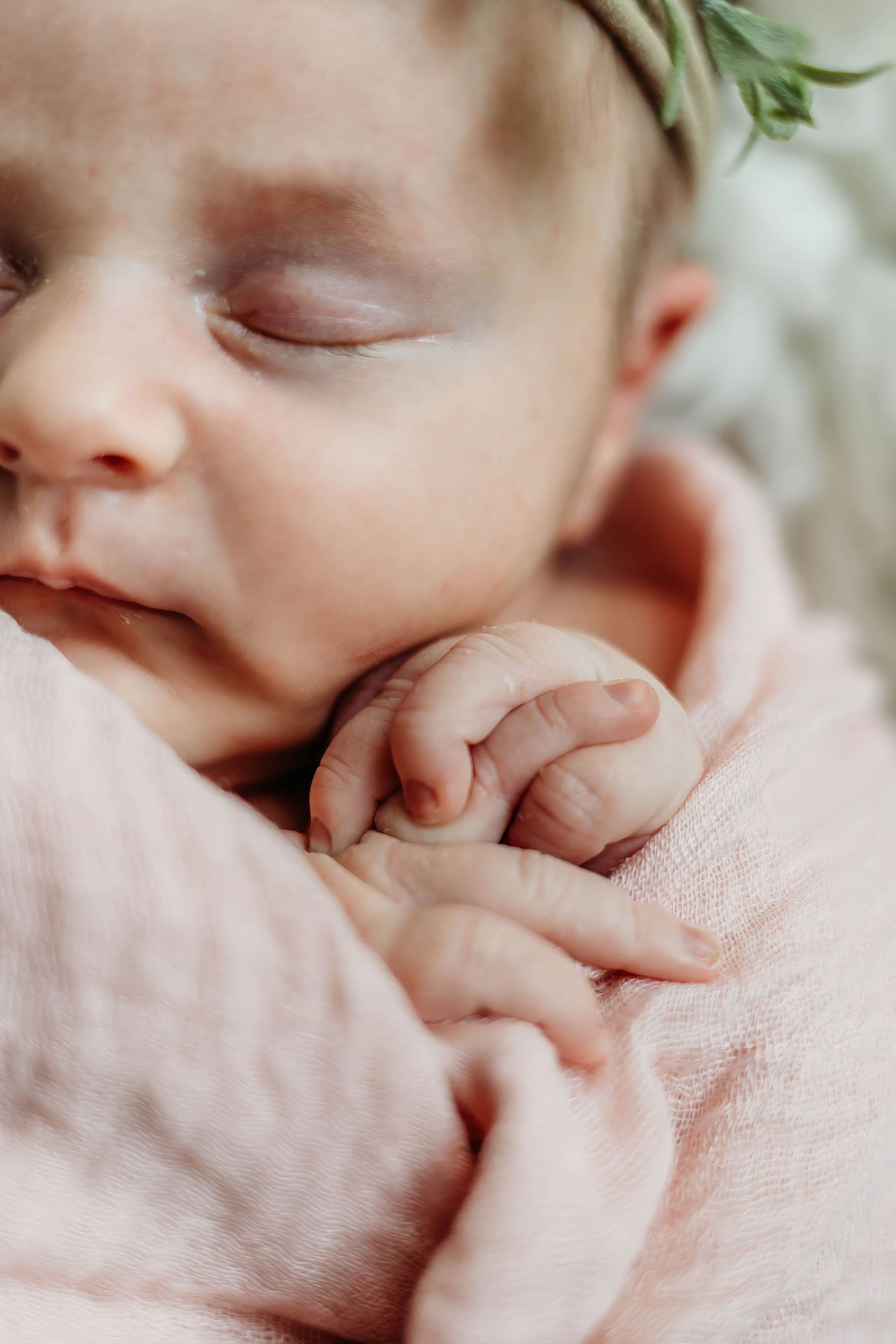 Close-up of a sleeping newborn baby wrapped in a soft pink blanket wearing a delicate green floral headband.