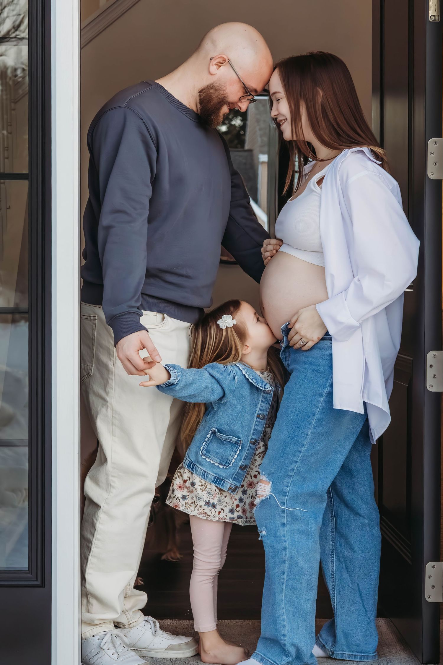 Family moment with bald father, pregnant mother, and young daughter standing in a doorway together.