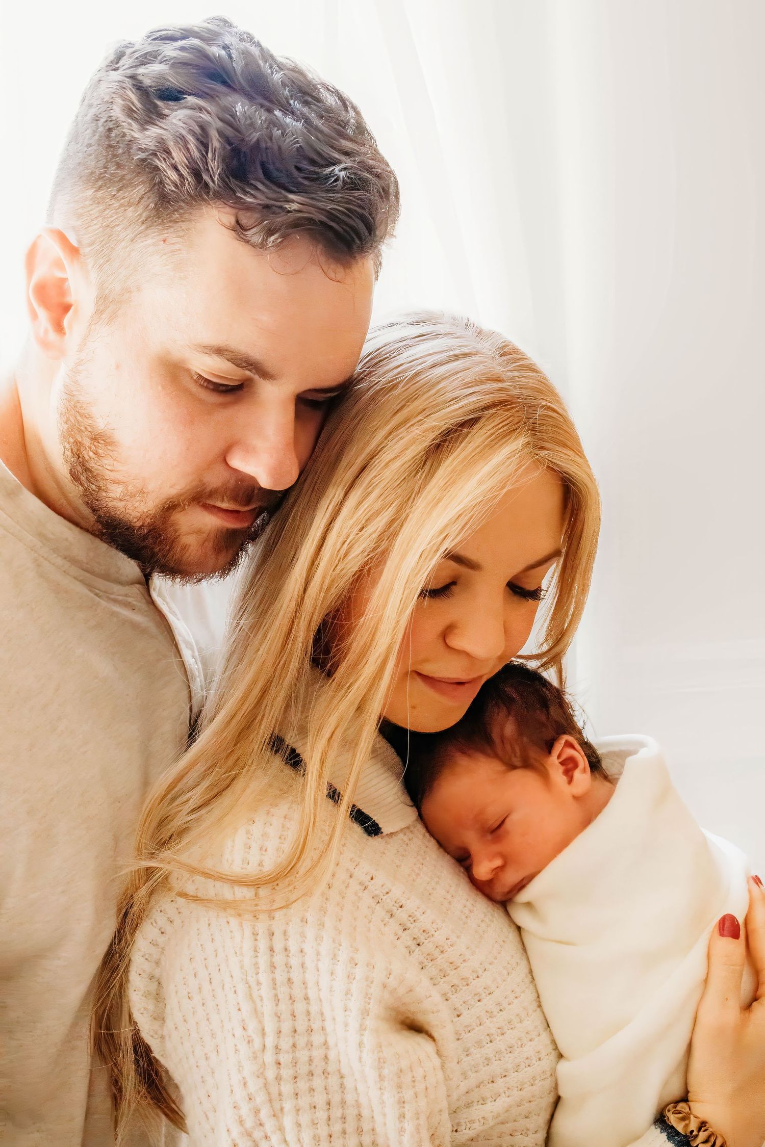 New parents tenderly gaze at their sleeping newborn baby wrapped in a white blanket near a window.