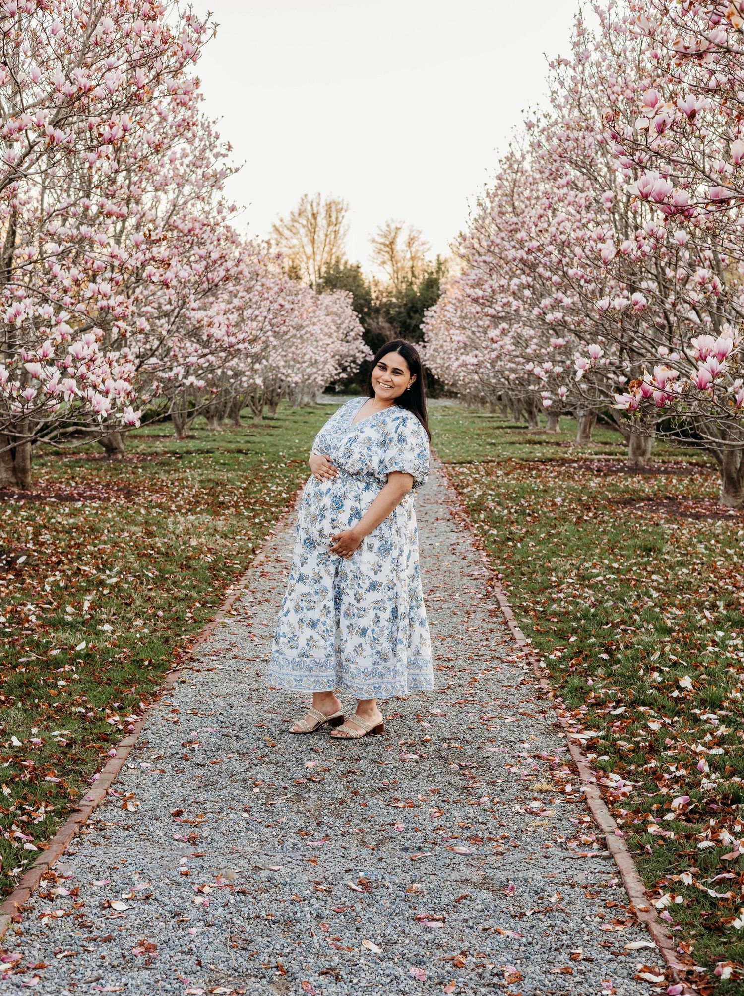 Pregnant woman in a floral blue dress poses among blooming pink magnolia trees in a garden path.