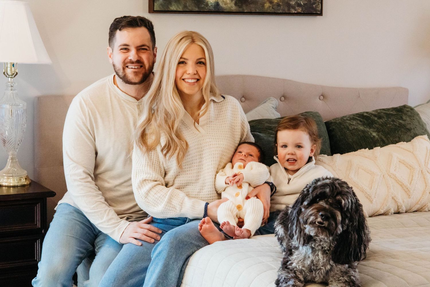 Happy family with newborn baby, toddler, and fluffy dog pose together on a bed for lifestyle photos.