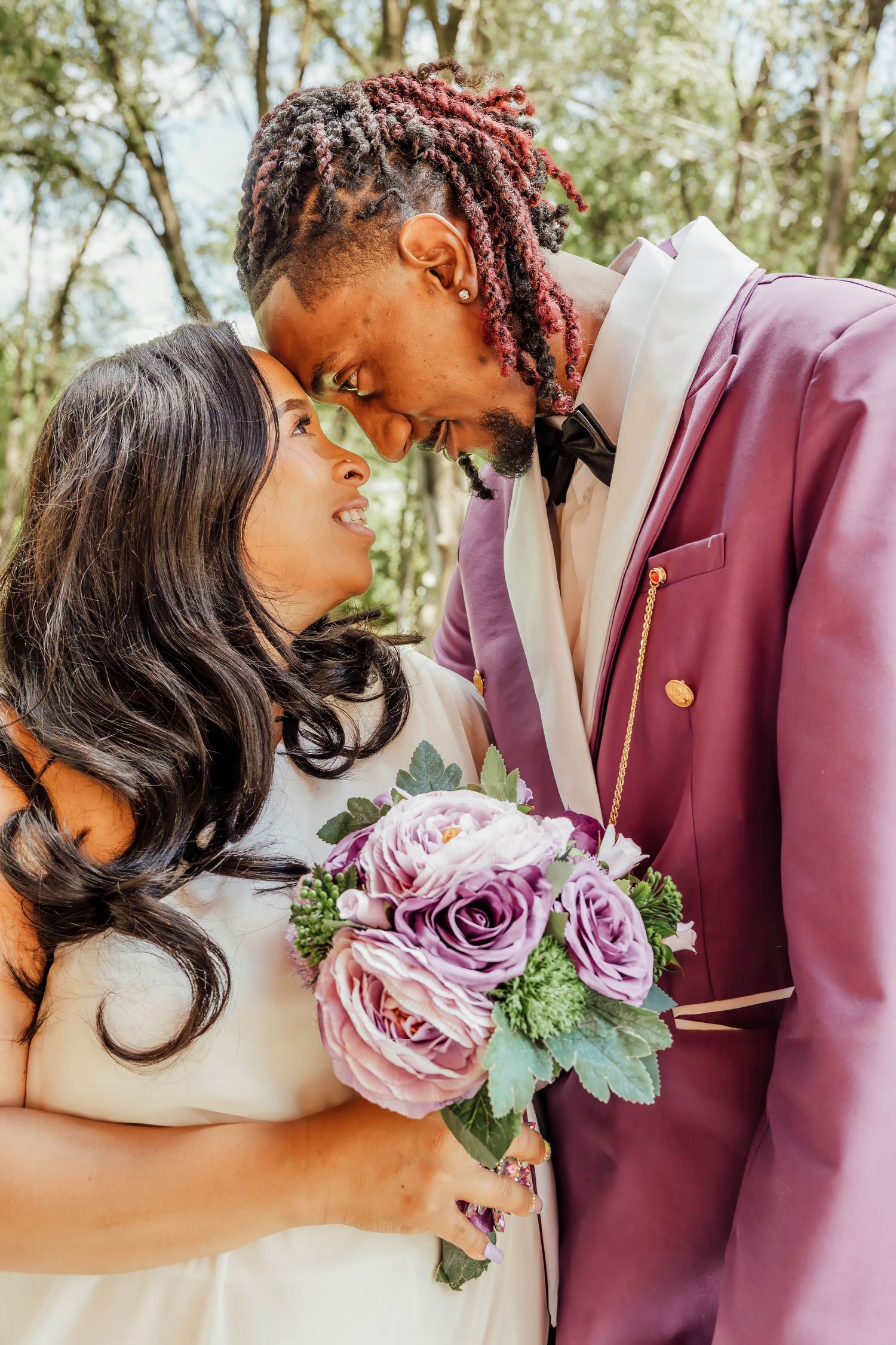 Newlywed couple in wedding attire touching foreheads, groom in purple suit holding bride with bouquet.