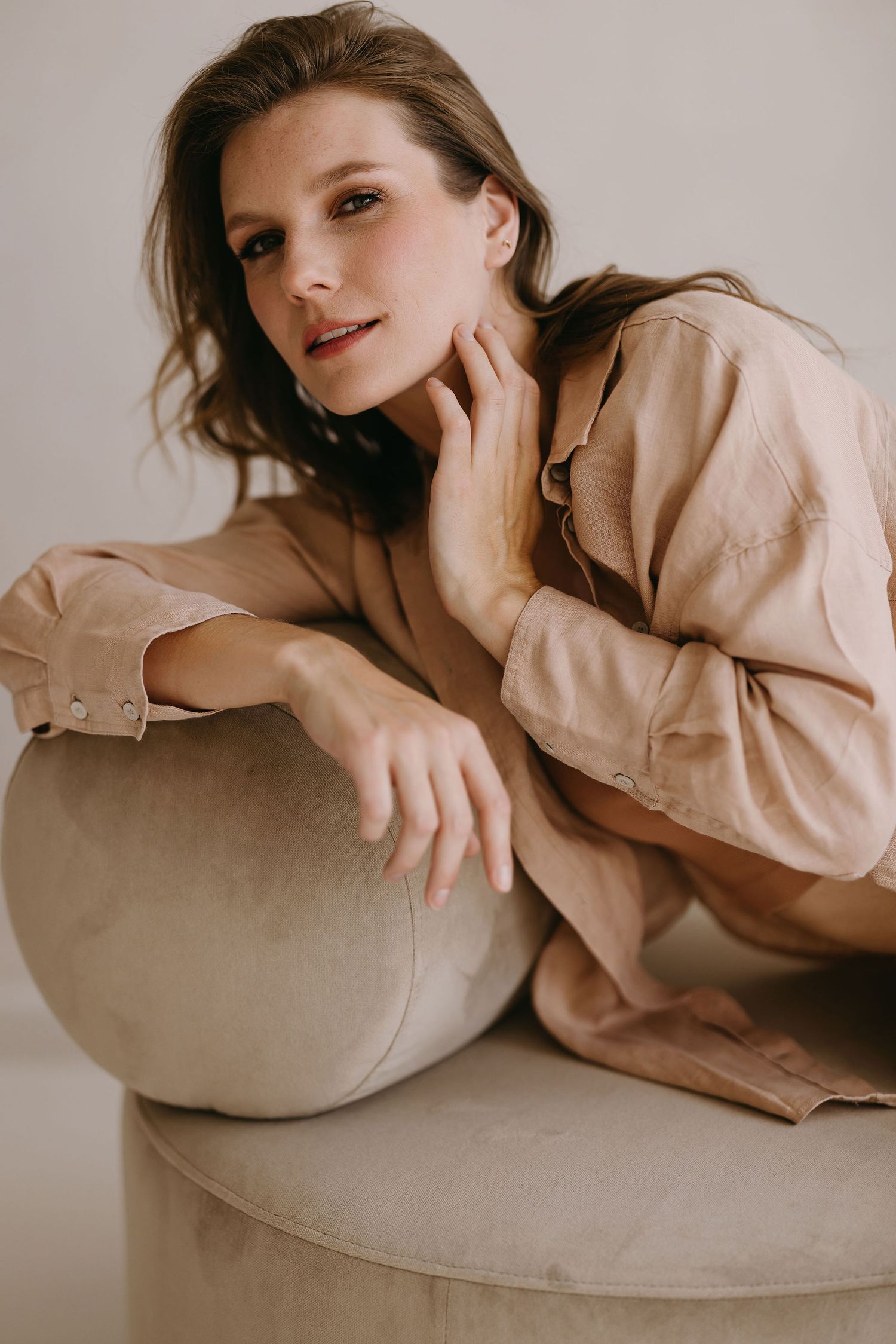 Fashion model in beige button-down shirt poses elegantly on cream-colored furniture in soft lighting.