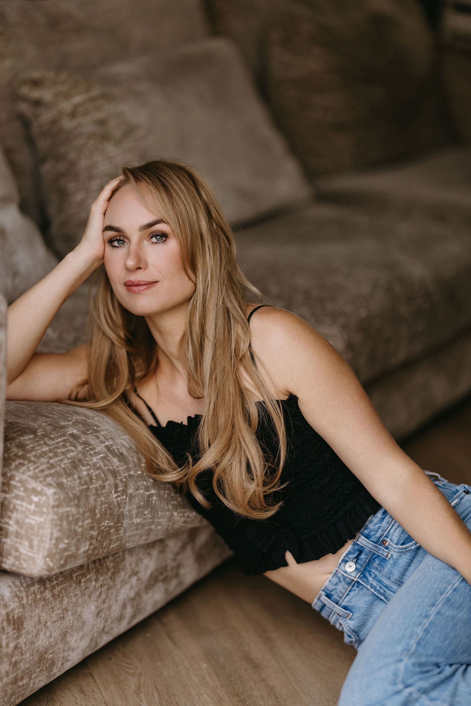 Casual indoor portrait featuring blonde hair, black top and denim in relaxed couch setting.