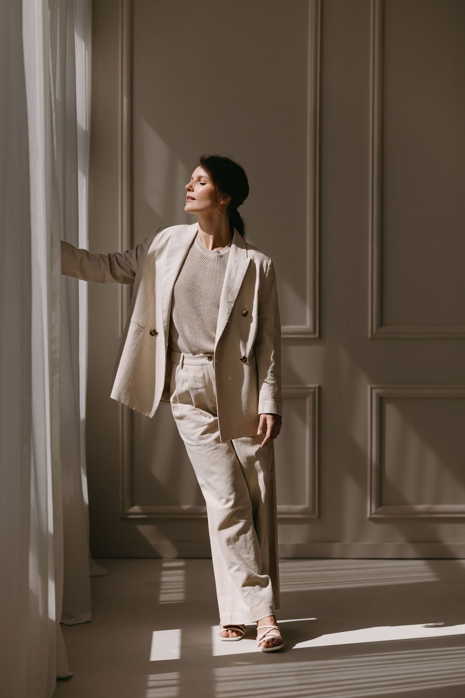 Model wearing cream colored suit poses by window with natural light casting dramatic shadows.