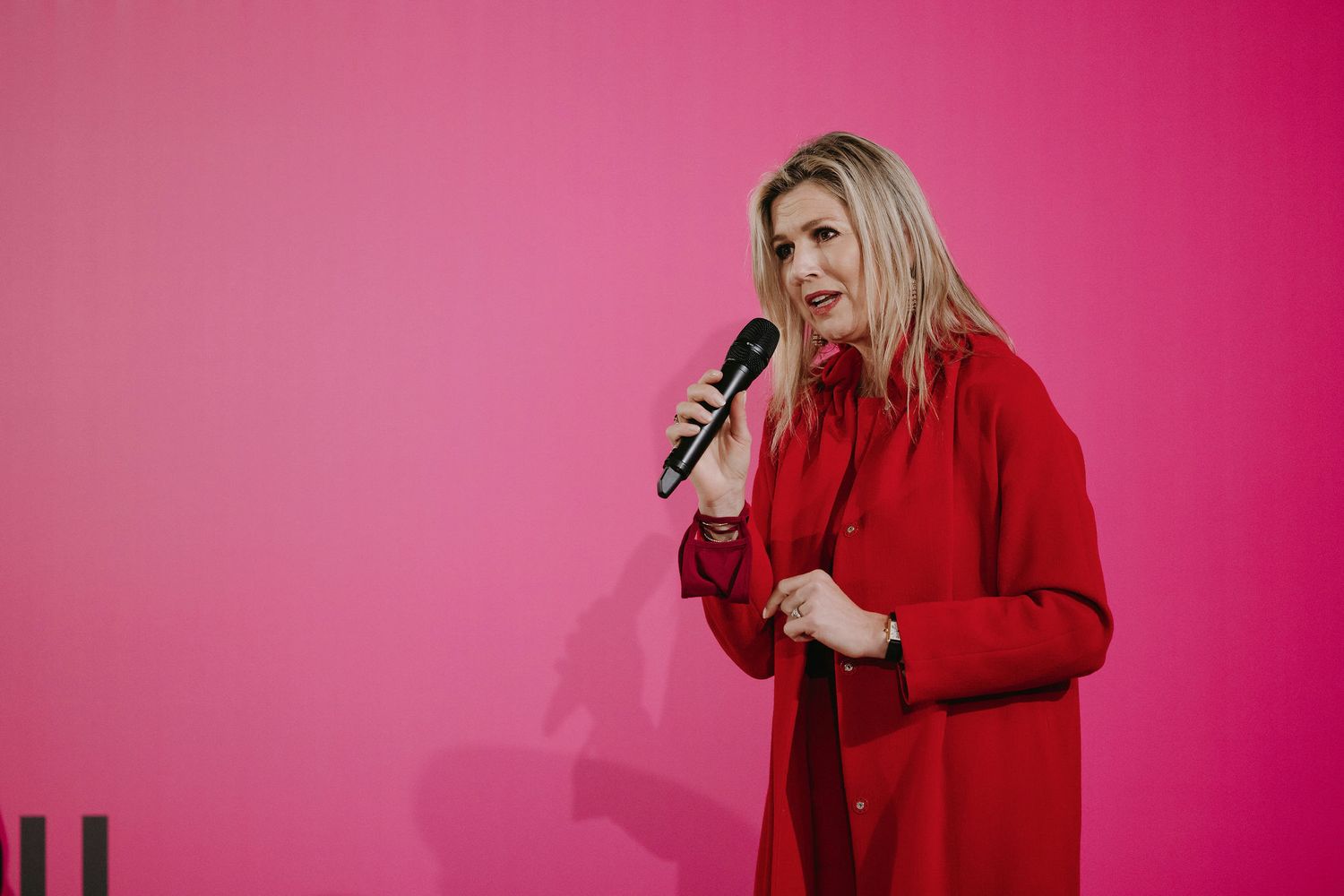 A speaker in a red coat delivers a presentation against a vibrant pink backdrop during an indoor event.