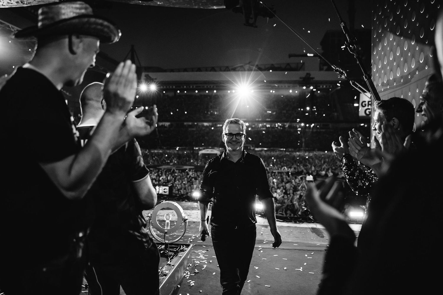 Performers celebrate on stage at a concert venue with confetti and bright lights.