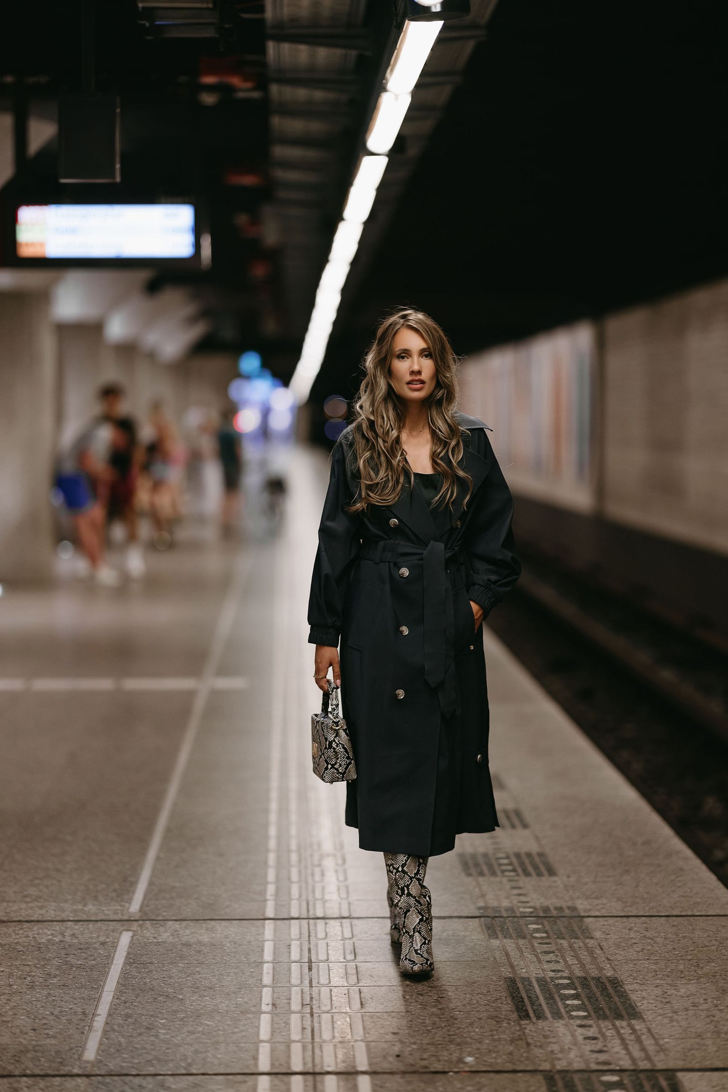 Fashionable figure in black trench coat and snakeskin boots walking through subway station.