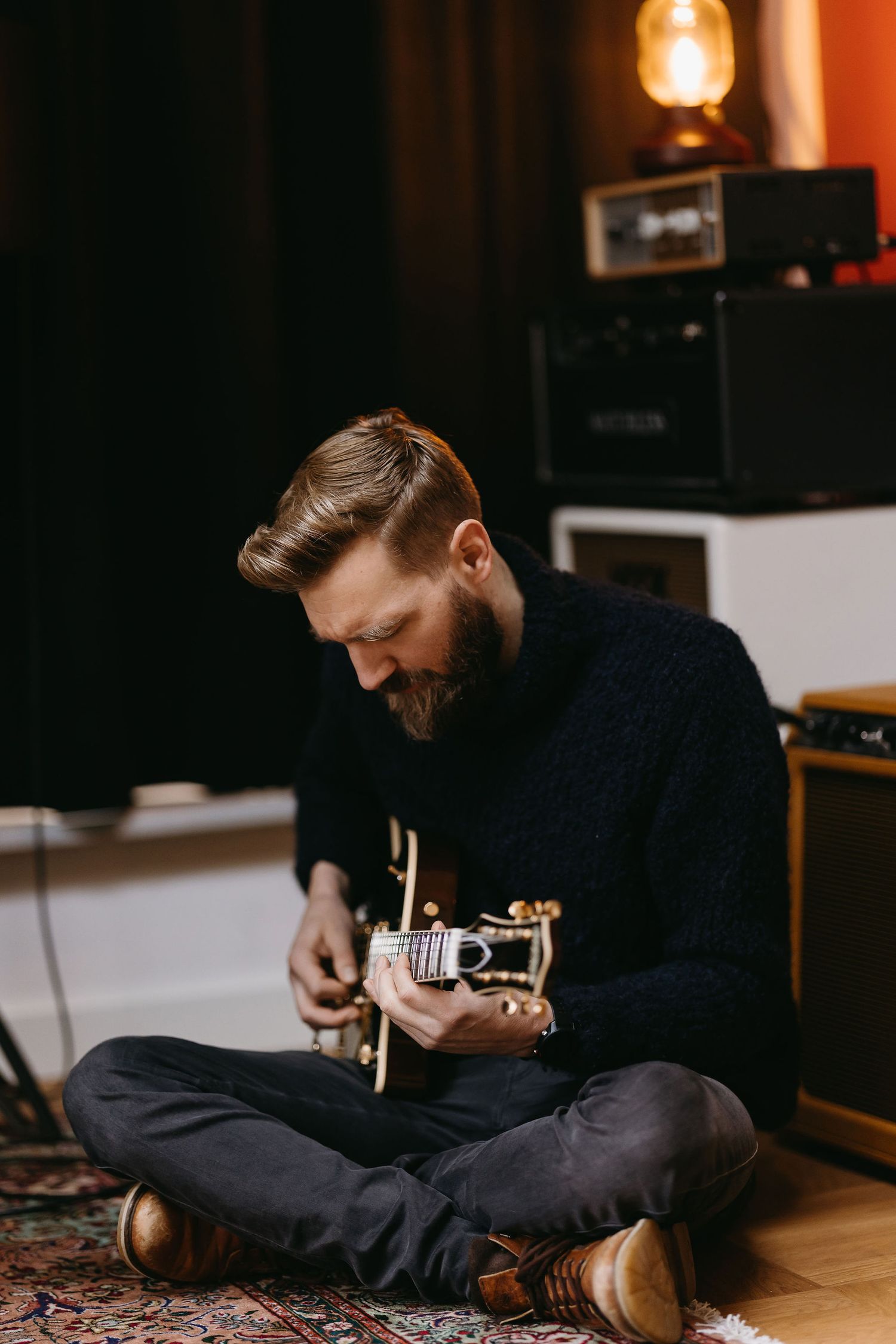 Musician playing acoustic guitar while sitting cross-legged on patterned rug.