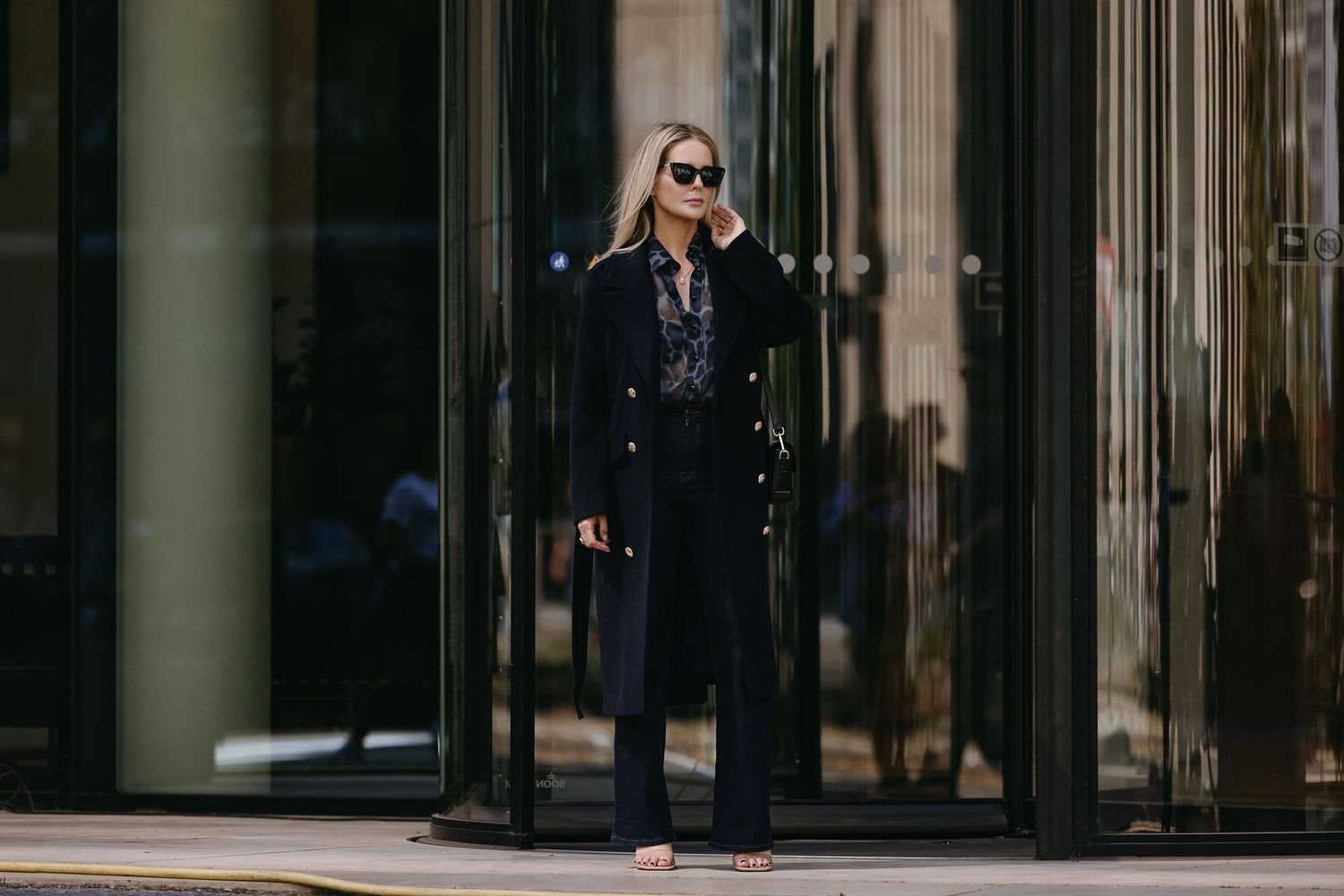 Fashionable street style featuring long black coat and sunglasses in urban setting.