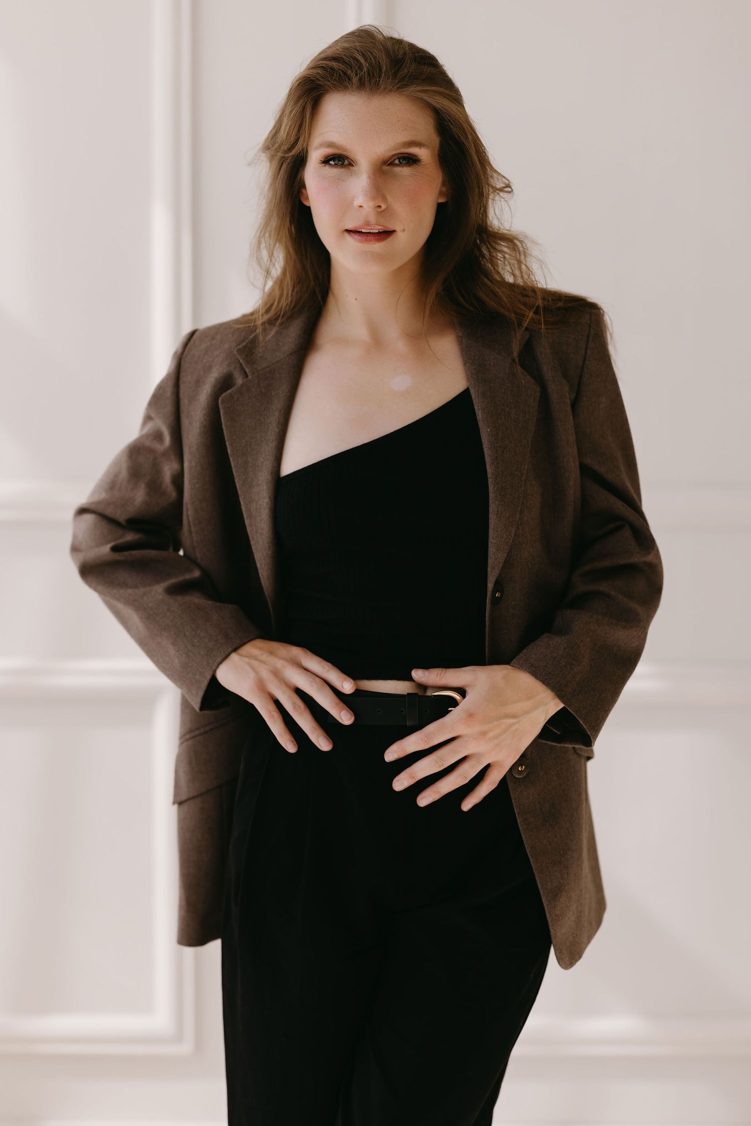 Fashion portrait featuring brown blazer over black dress in minimalist white interior.