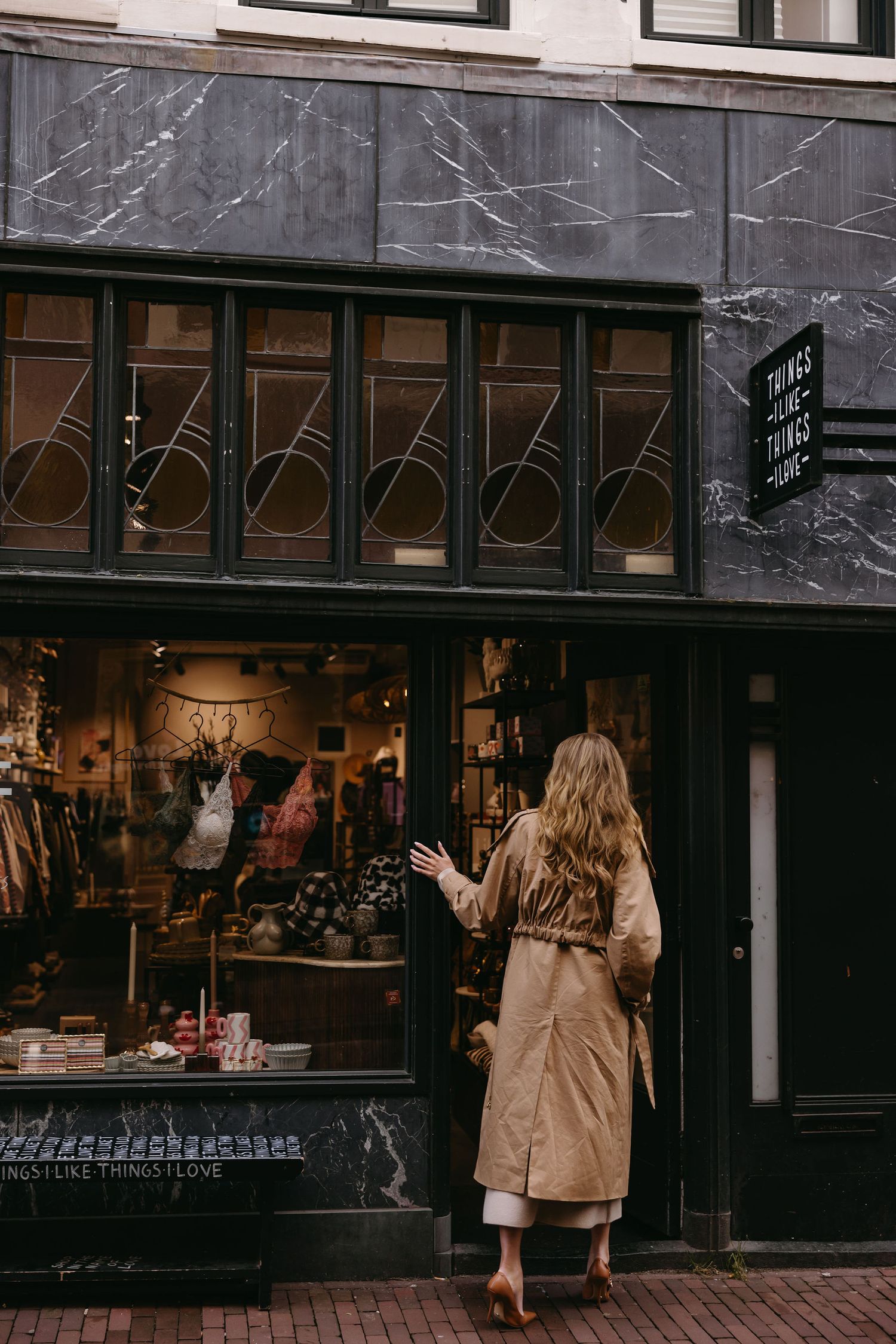 Woman in beige coat looking at vintage storefront display with marble exterior.