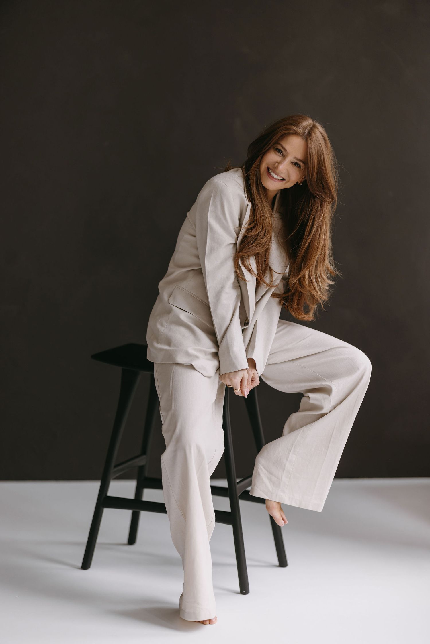 Casual fashion portrait of model in cream suit sitting playfully on black stool.