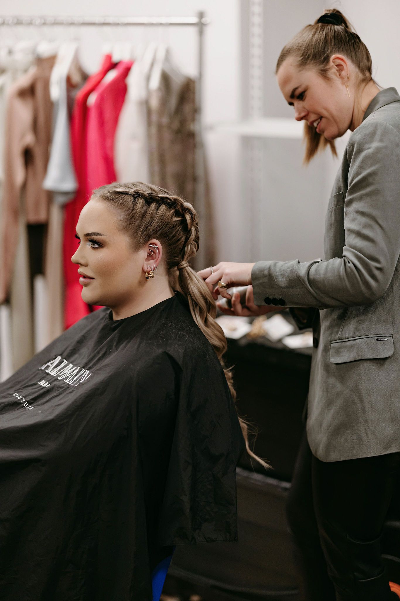Hair stylist creating an intricate braided hairstyle in a salon with clothing racks visible in background.