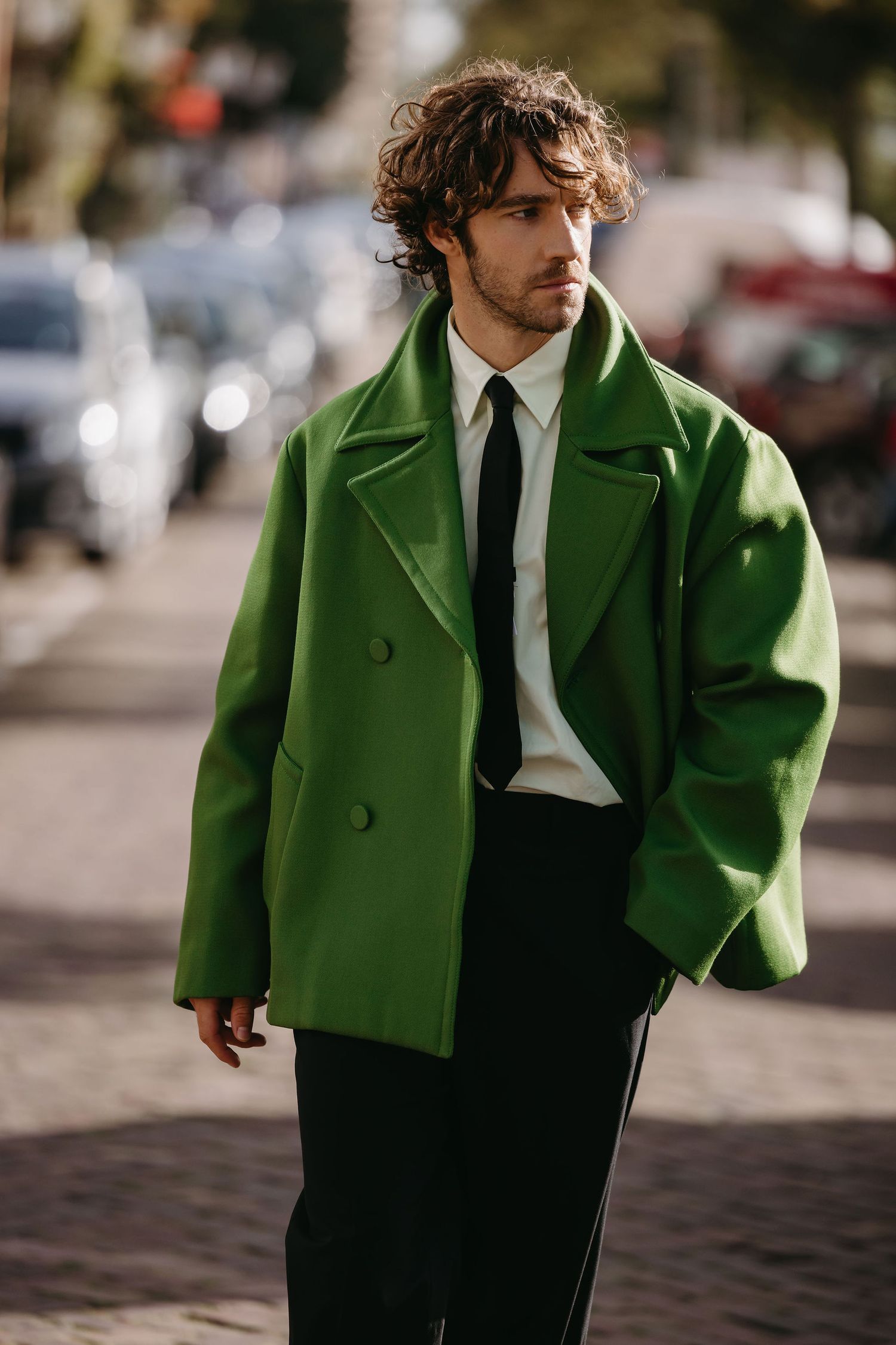 Model wearing vibrant green overcoat with white shirt and tie on city street.
