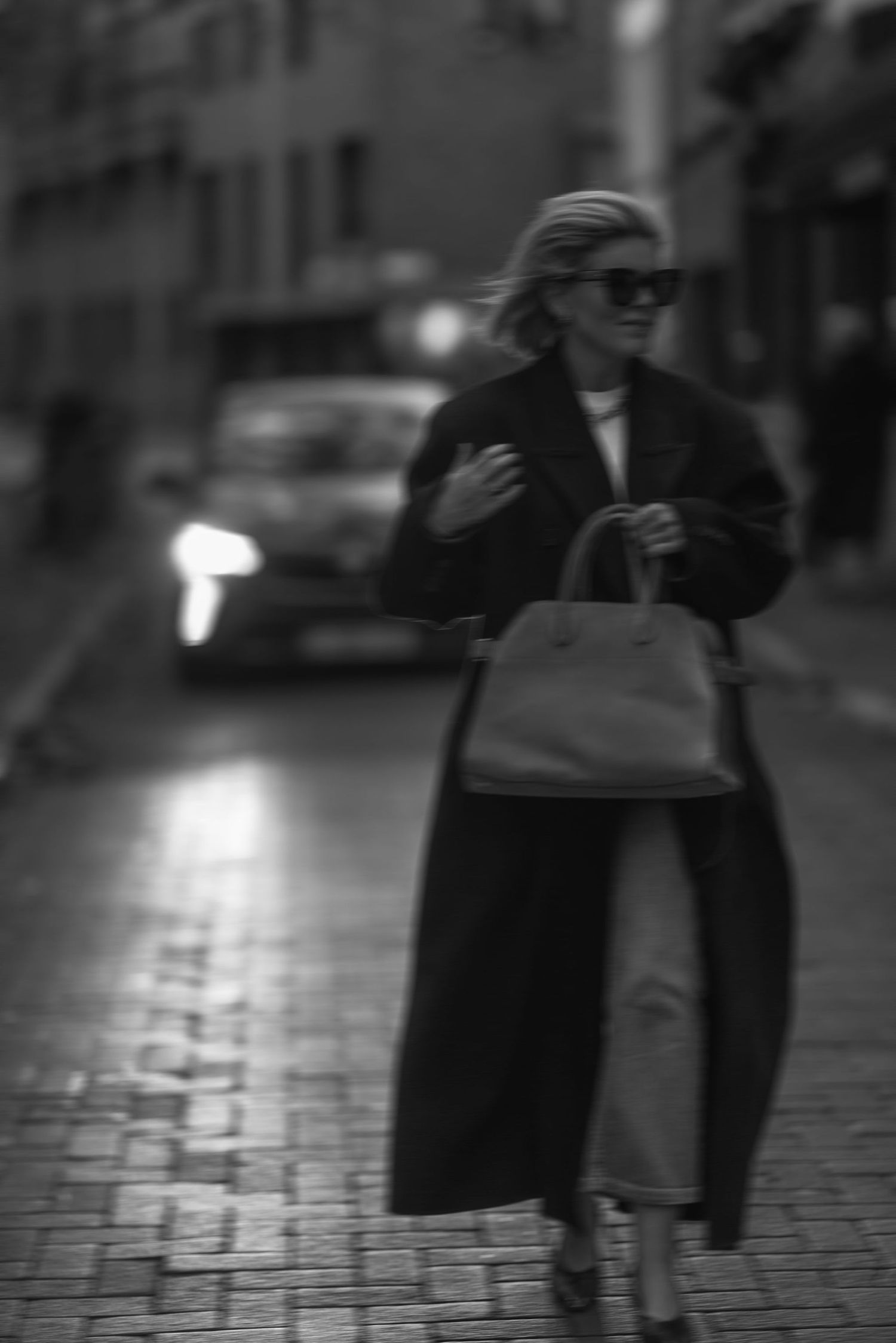 Moody street style photograph featuring elegant black coat and handbag on rainy cobblestone street.