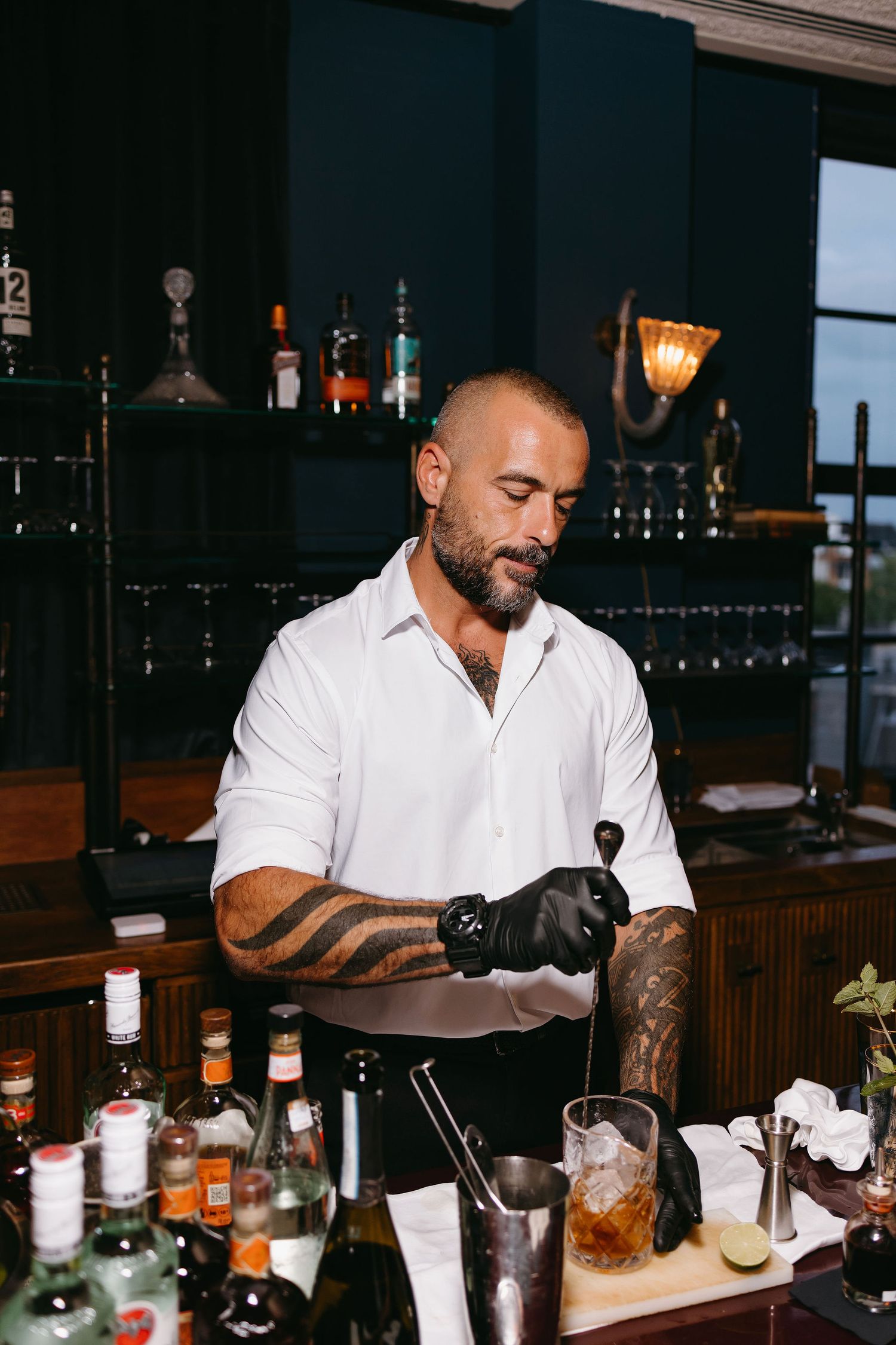 Tattooed bartender in white shirt preparing cocktails at well-stocked bar.
