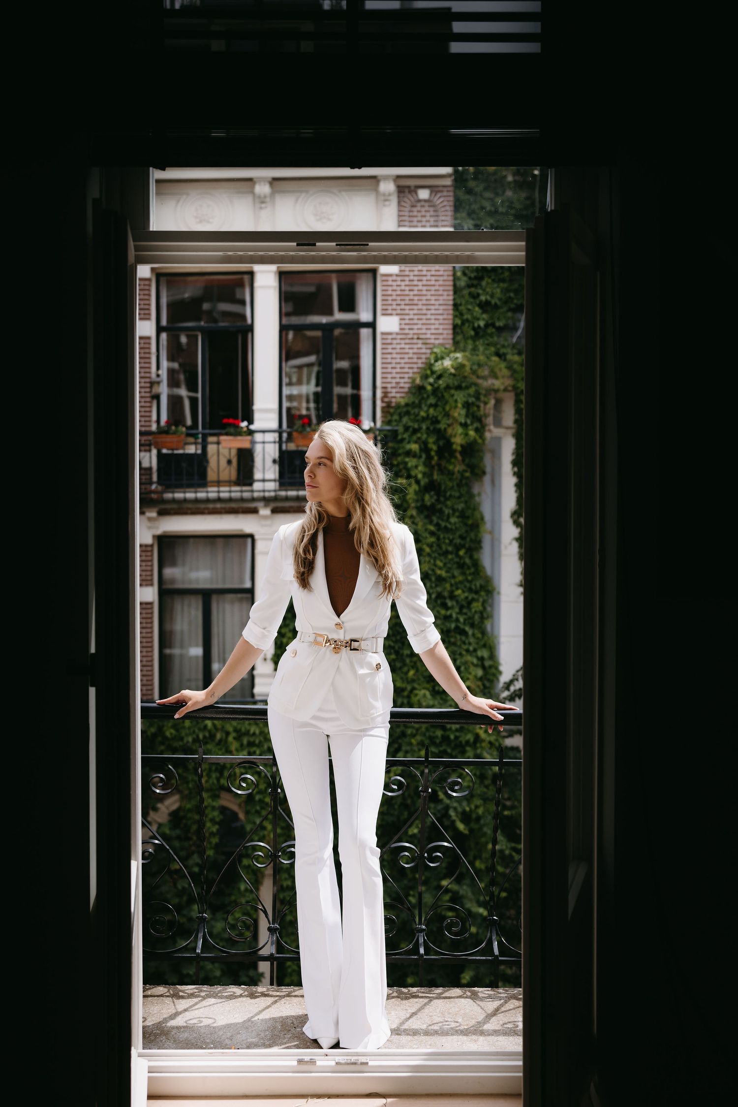 Person in all-white outfit standing on balcony against urban backdrop.