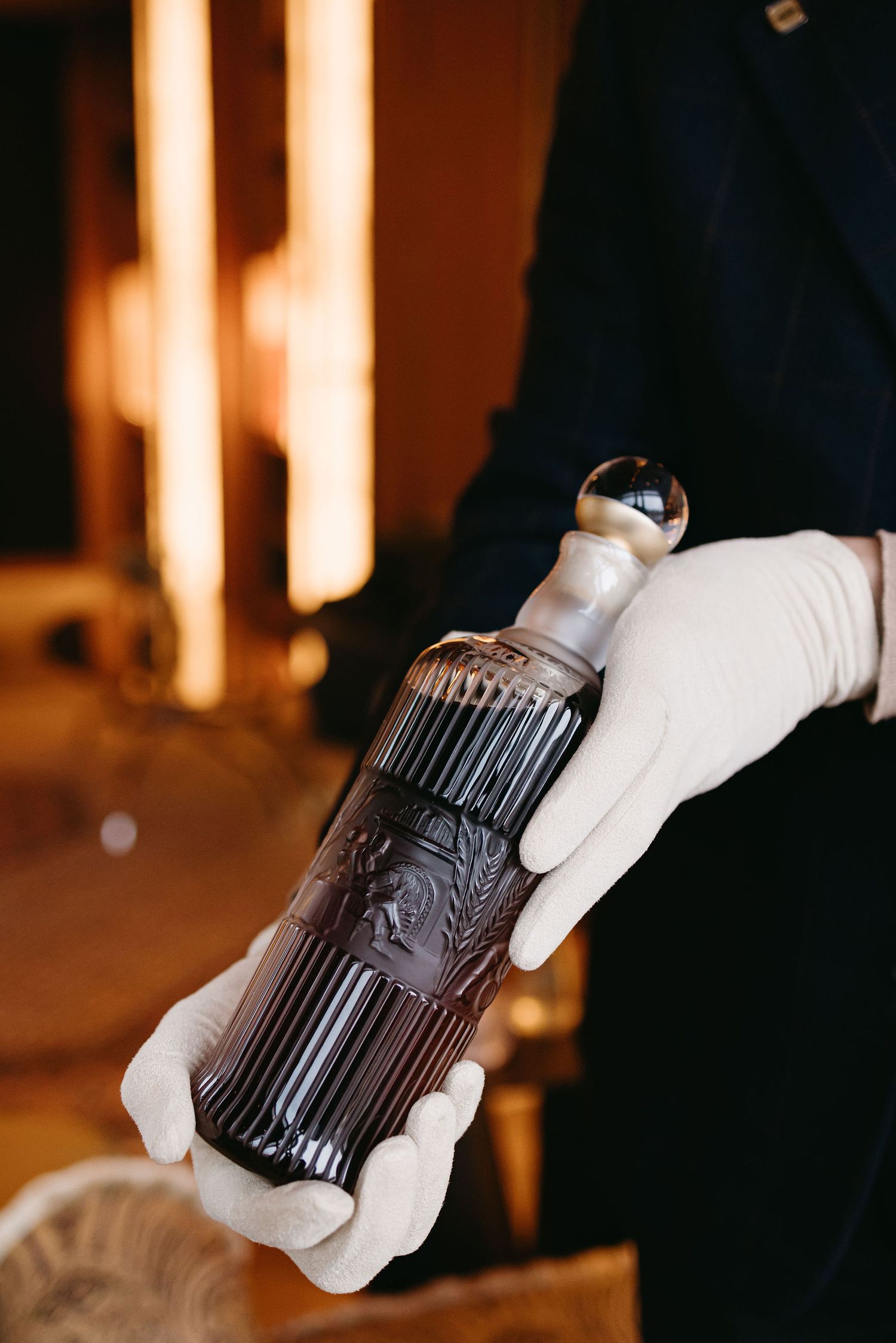 Hands wearing white gloves hold an ornate dark glass bottle against a warm background.